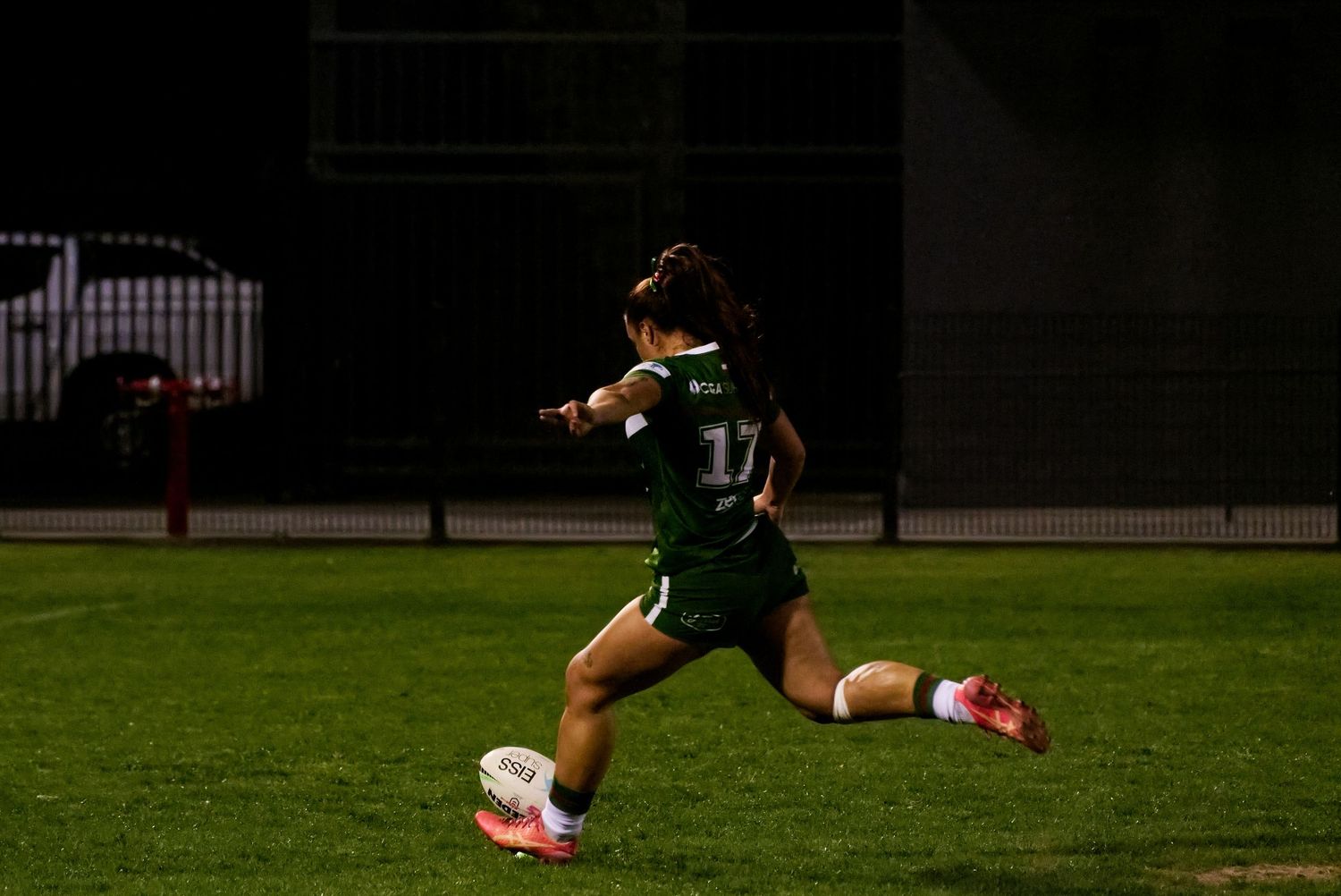 Female rugby league player wearing green jersey number 11 kicking ball during night match on floodlit field.