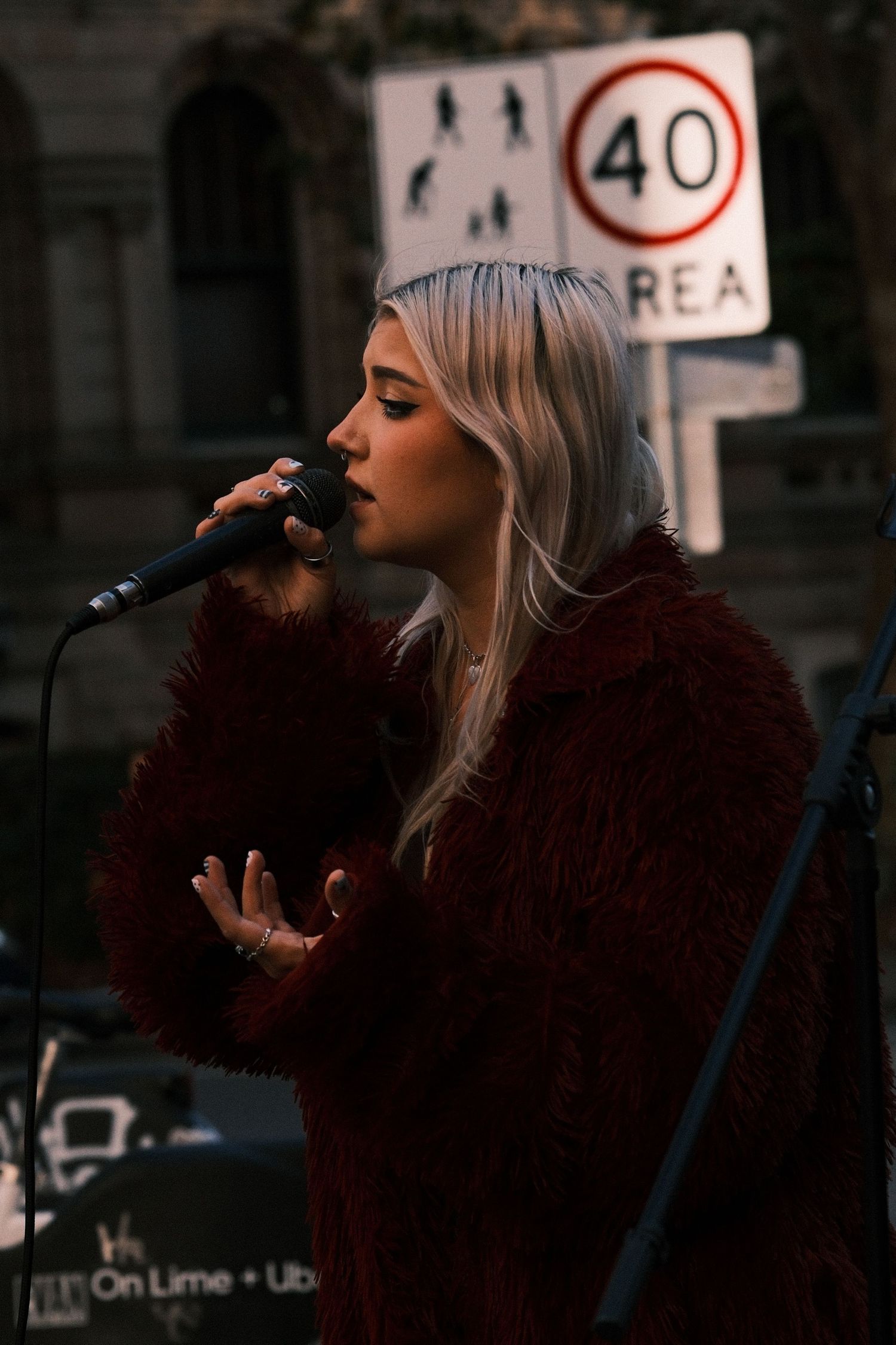 Blonde woman in red fur coat singing into microphone outdoors near a 40km/h speed zone sign.