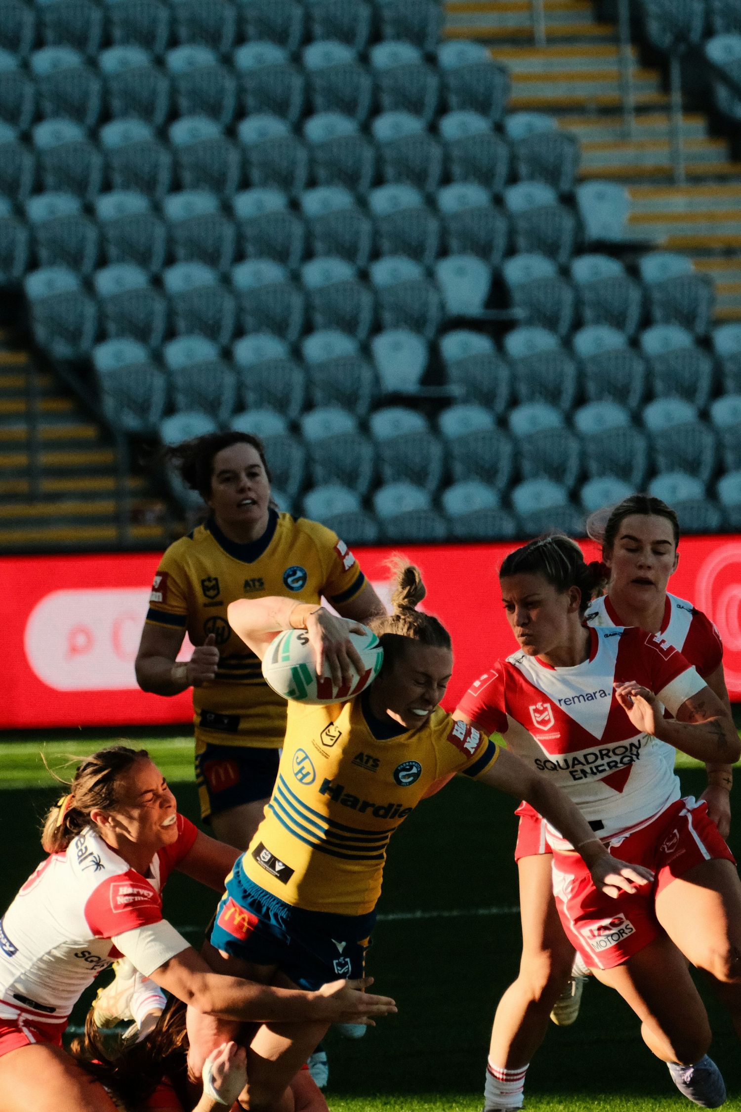 Women's rugby league match with players in yellow and red jerseys competing for the ball in a stadium.