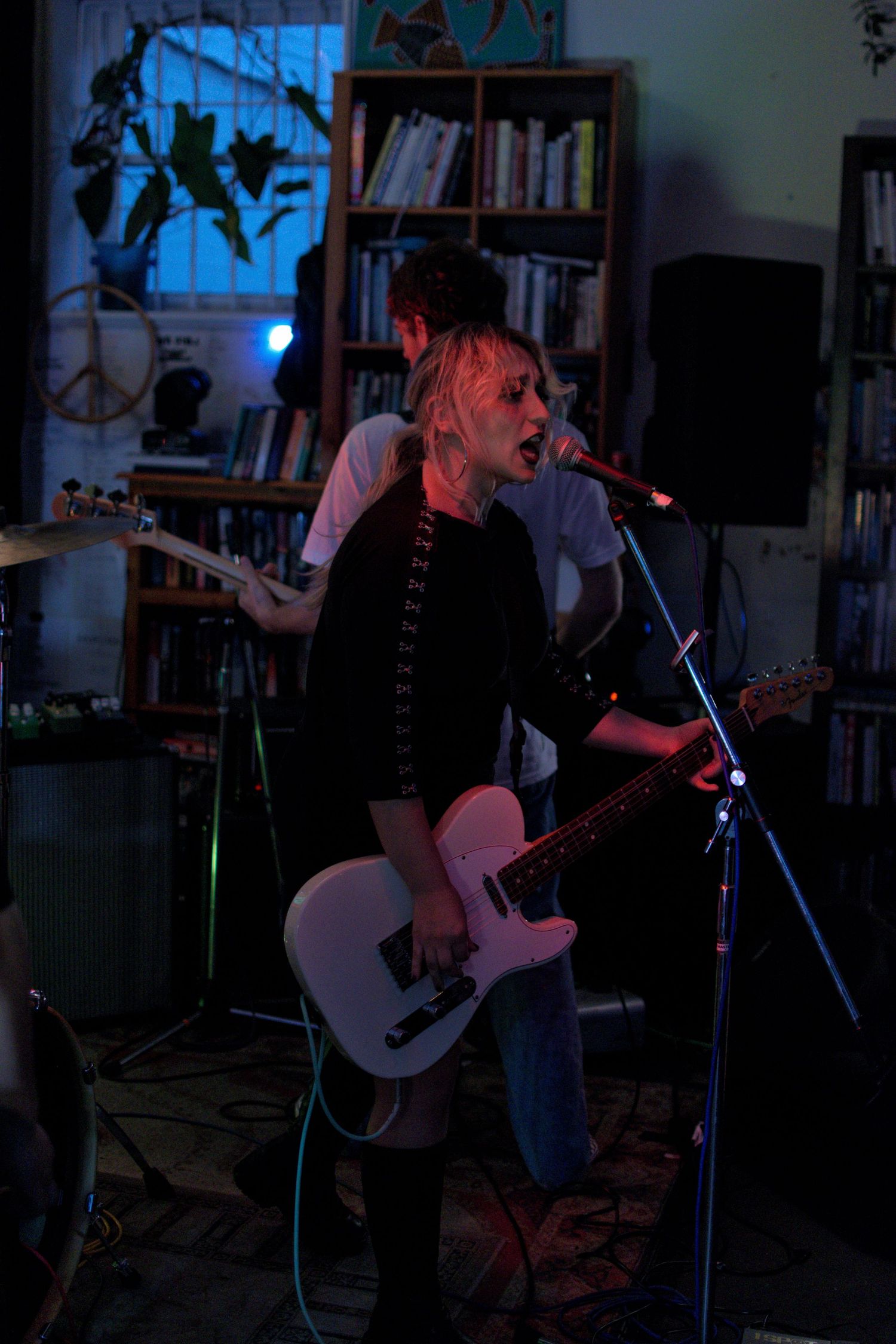A female guitarist with blonde hair singing into a microphone while playing a white electric guitar at an indoor show.