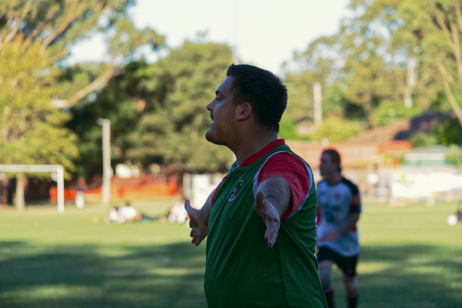 A soccer coach in green and red uniform gestures on the sideline during an outdoor match.