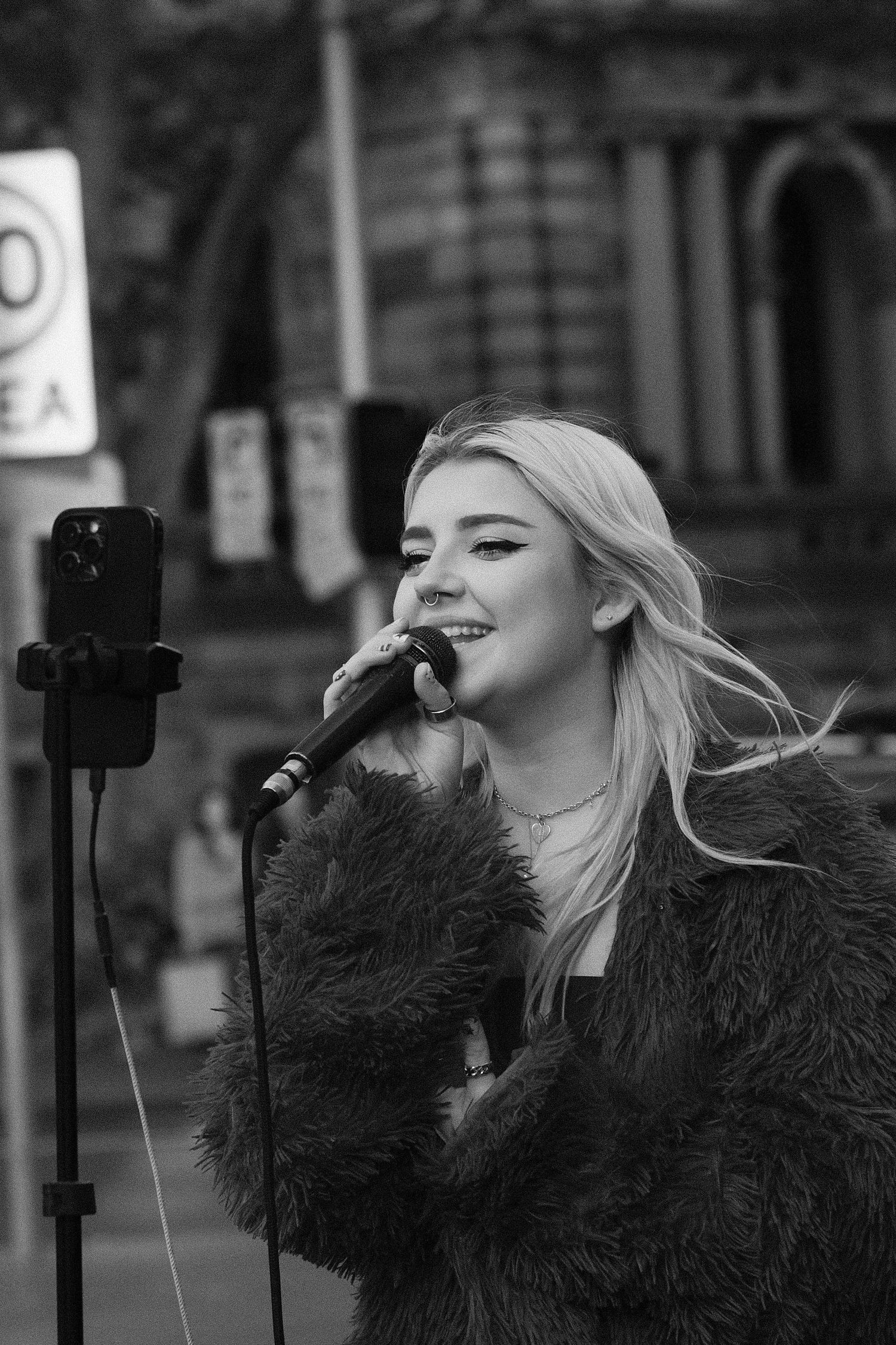 Young blonde woman singing into a microphone outdoors, wearing a fluffy coat, with a phone on a stand beside her.