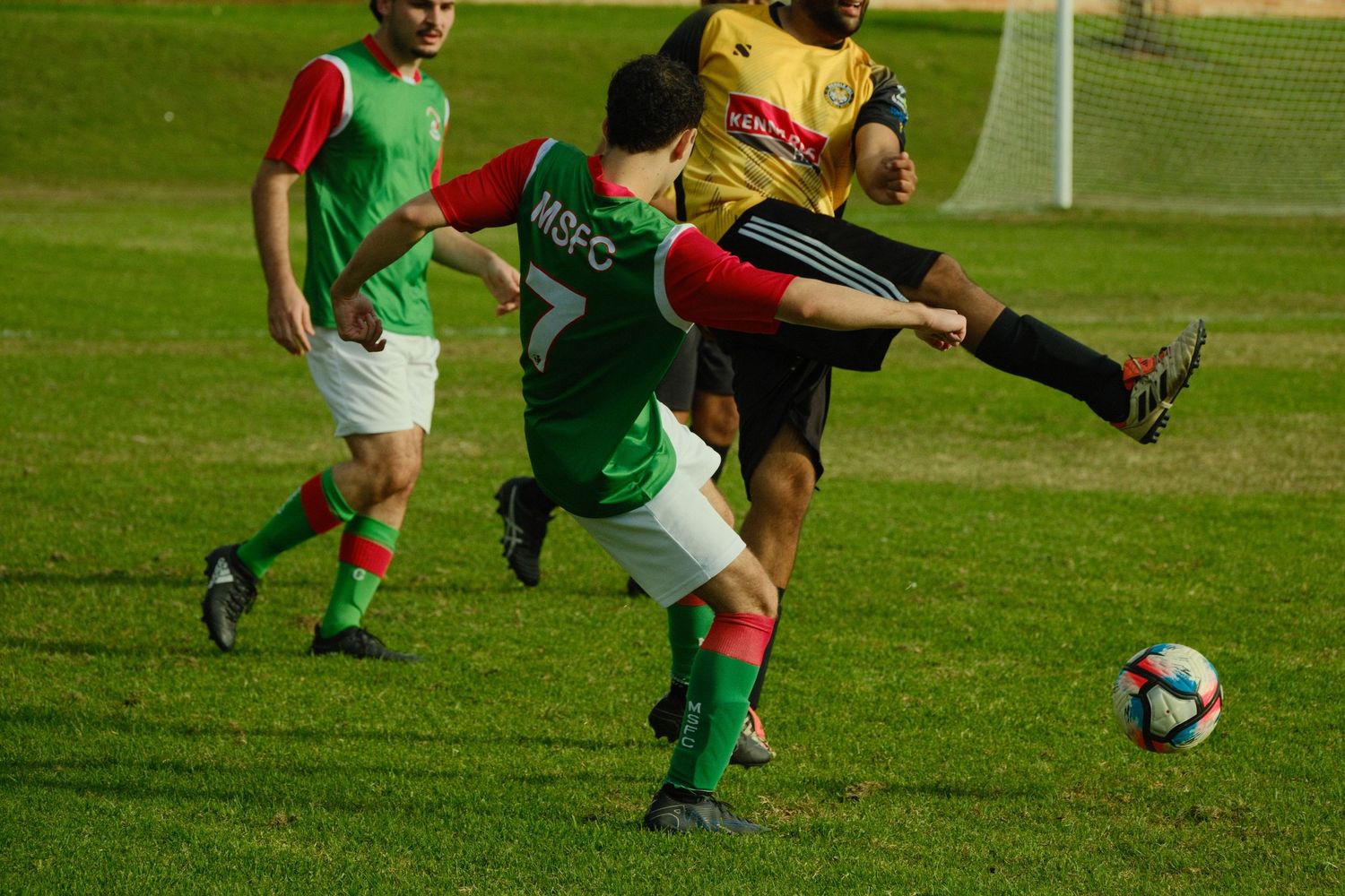 Two soccer players compete for the ball during a match, one in green and red kit, the other in yellow and black.