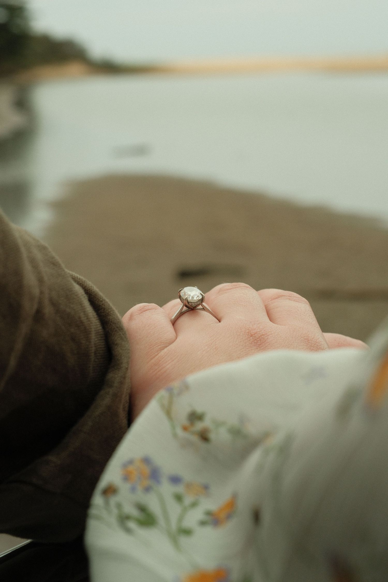 Close-up of an engagement ring on a woman's hand held by her partner near a sandy beach shoreline.