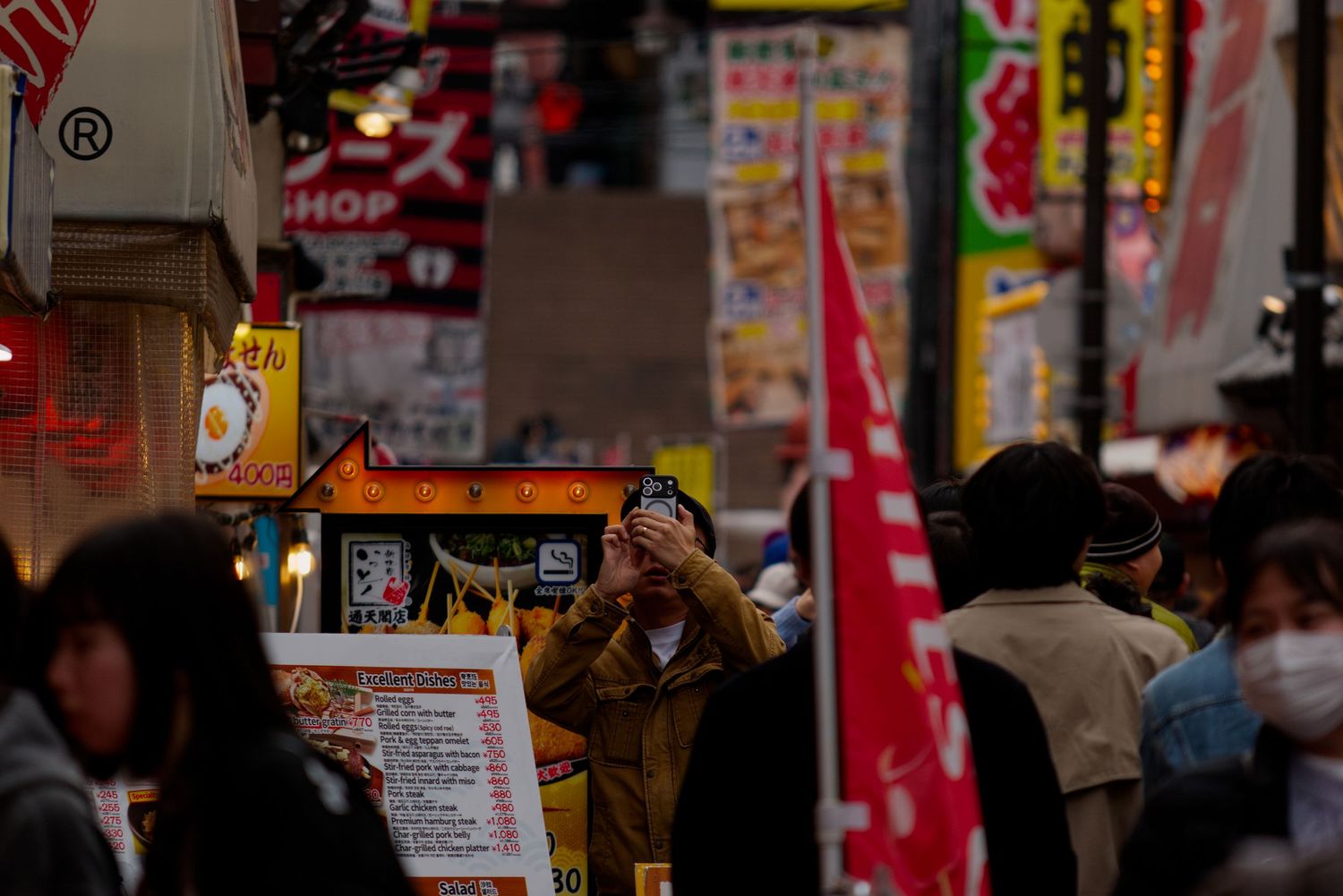 Busy Japanese street market with crowds of people walking past colorful signs and shops in a vibrant city district.