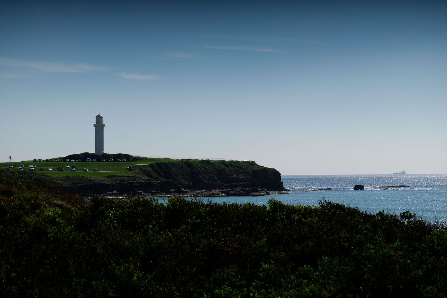 A lone lighthouse stands on a green coastal cliff overlooking a calm blue ocean under a clear sky.