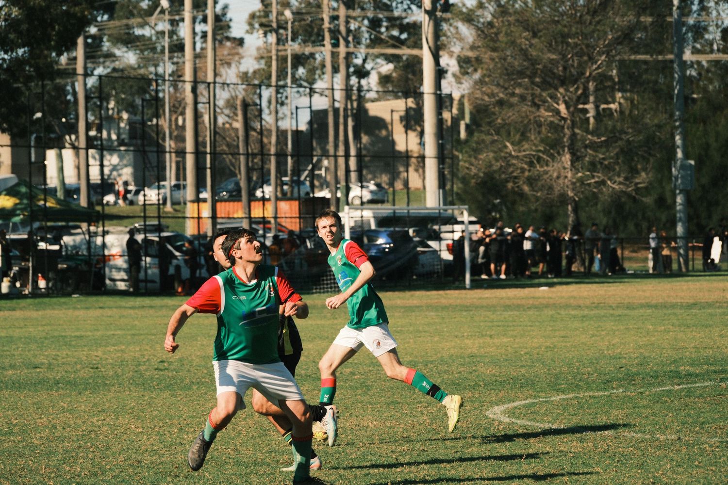 Two soccer players in green and red jerseys compete on a sunny grass field during an outdoor match.