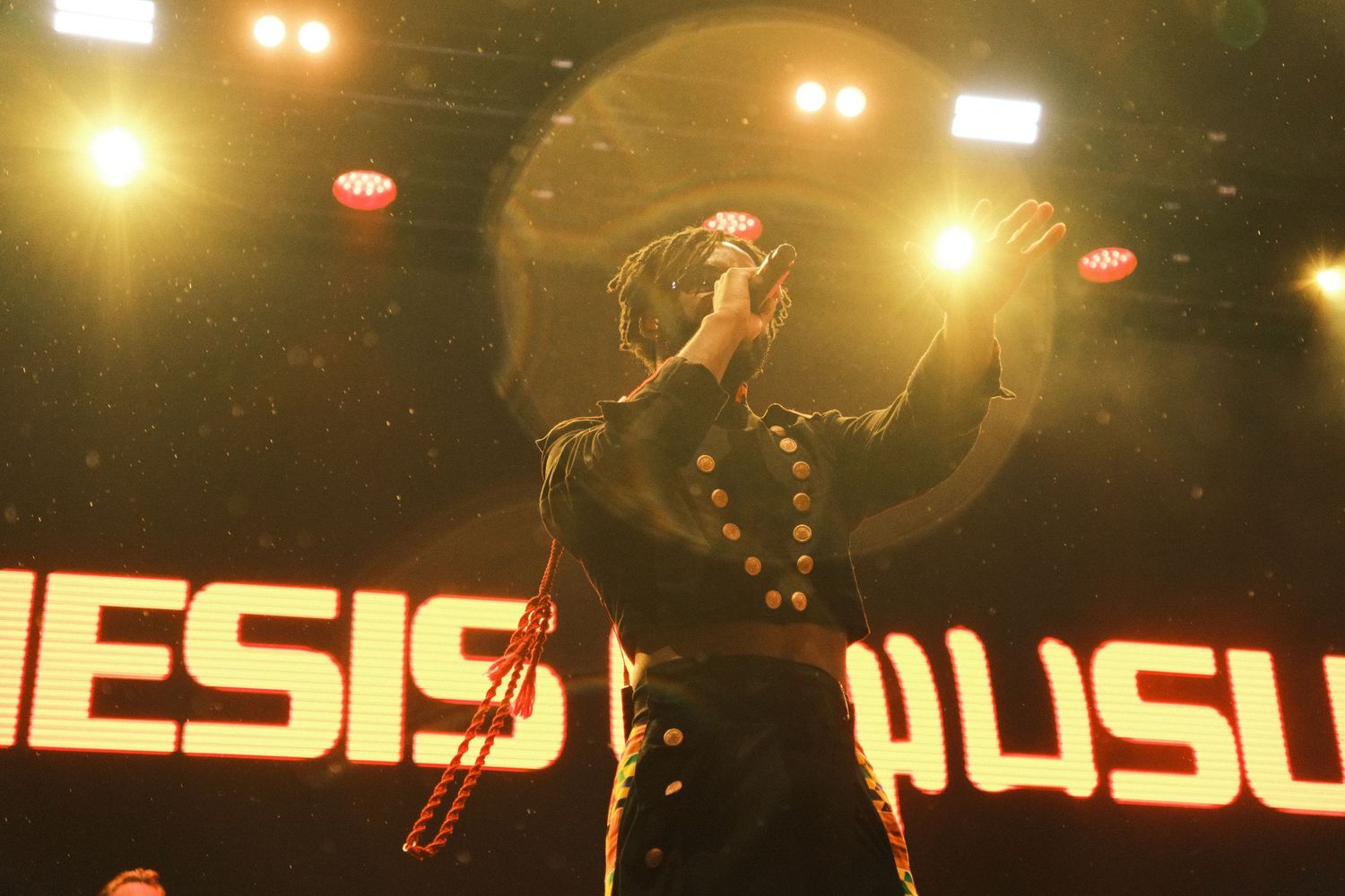 Singer performs on stage at Genesis Musus concert, backlit by golden spotlights with bold signage behind them.
