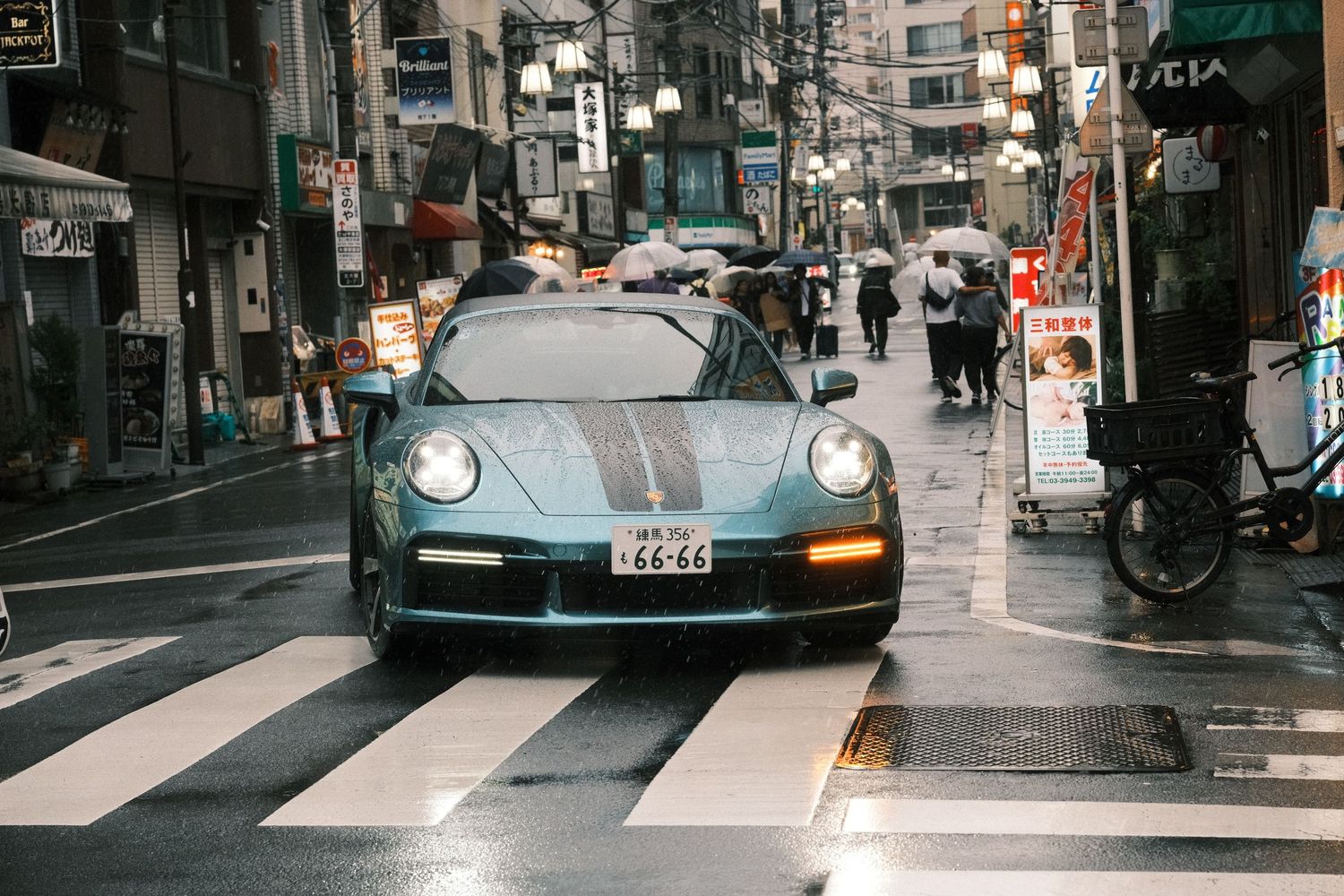 A green Porsche 911 drives through a rainy urban street in Japan with pedestrians and neon signs in the background.