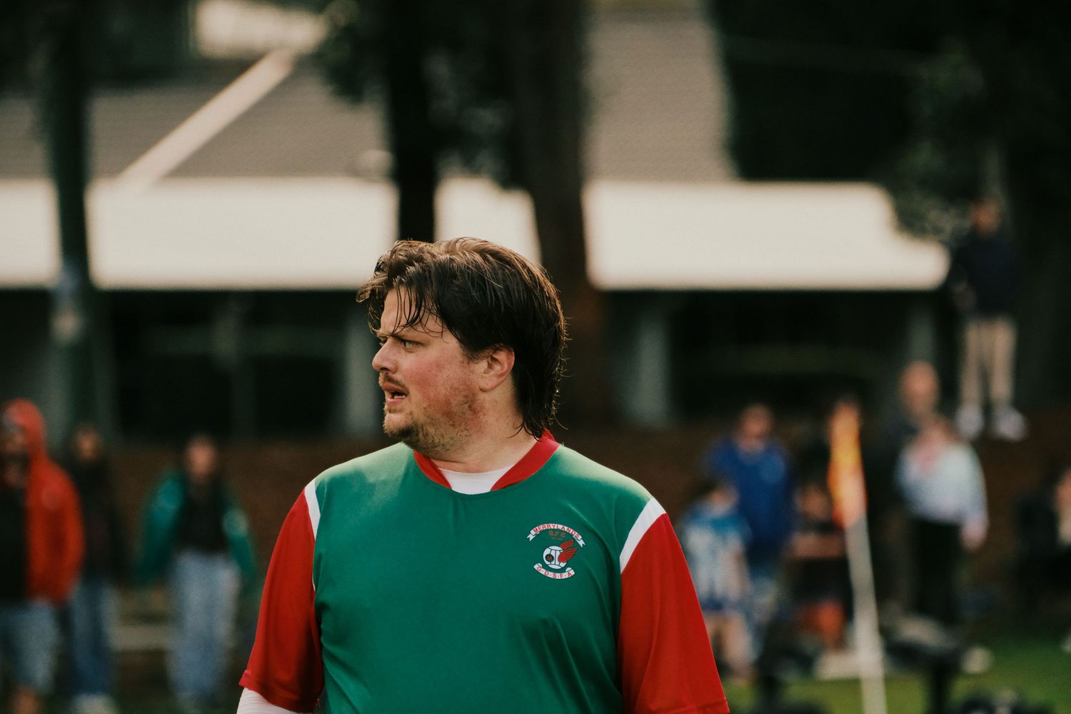Man in green and red soccer jersey looking sideways on a sports field with players in background.