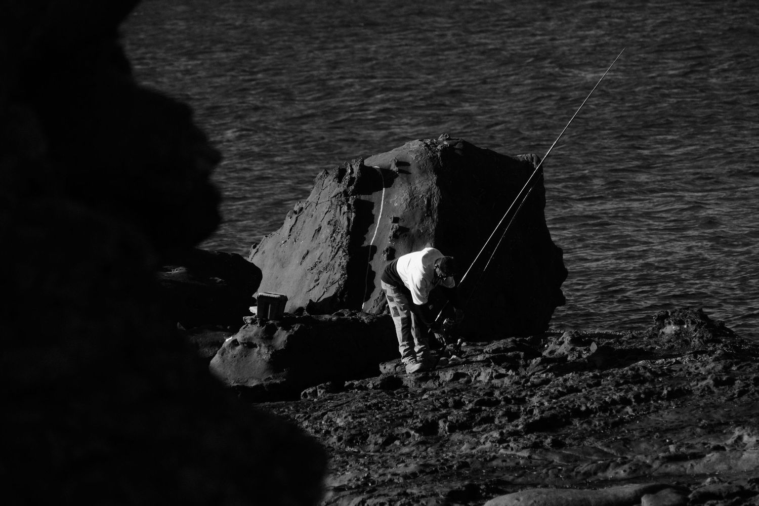 Black and white photo of a lone fisherman casting a line from rocky coastal cliffs beside dark ocean waters.