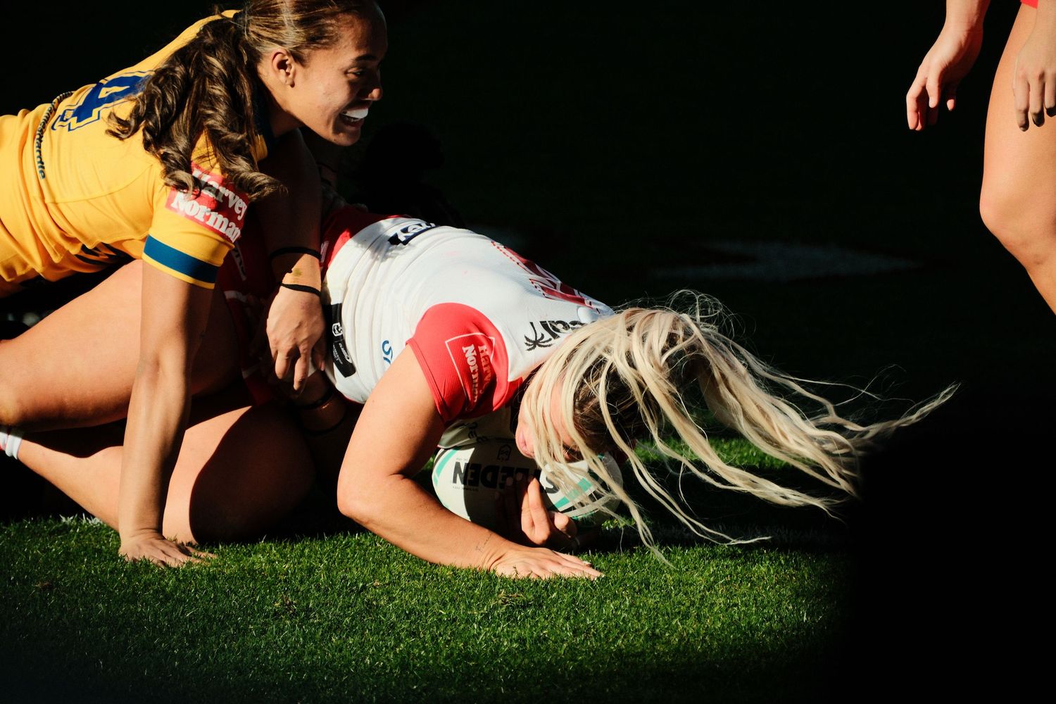 Female rugby players competing in a tackle during a women's rugby league match on a grass field.