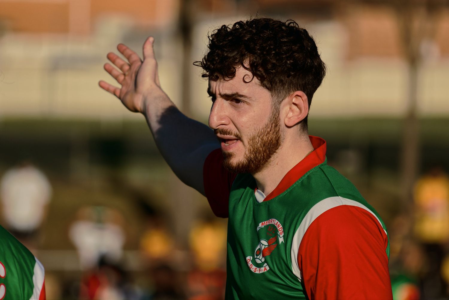 Young male soccer player in green and red MSFC jersey gestures with raised hand on field.
