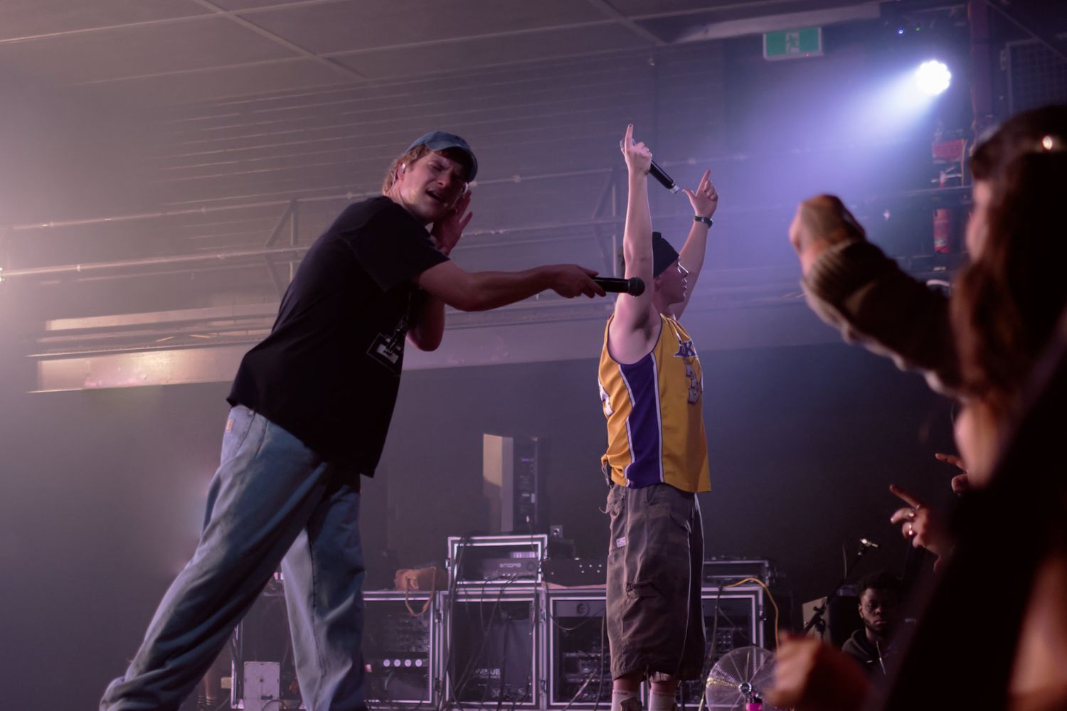 Two performers energize a crowd on a lit concert stage, one raising a microphone triumphantly under colorful spotlights.
