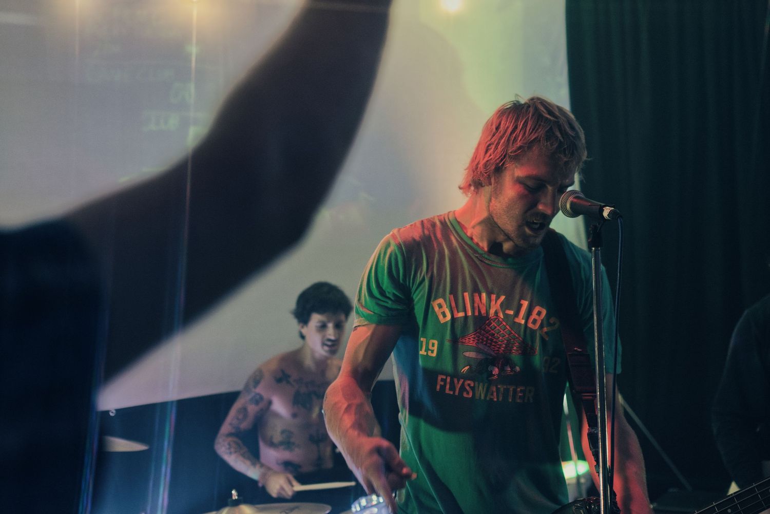 A tattooed male vocalist in a Blink-182 t-shirt performing on stage with a drummer visible in the background.