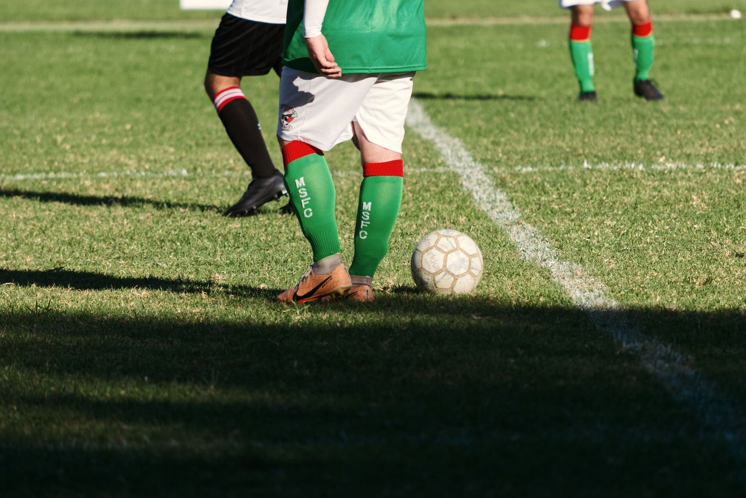 Close-up of soccer players competing for the ball near the sideline, wearing green and black socks.