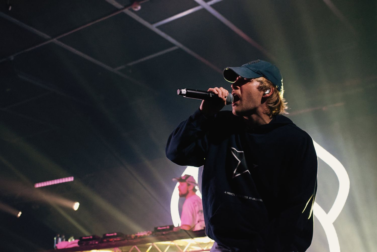 Hip-hop performer sings into mic on stage with DJ and neon lights in background.
