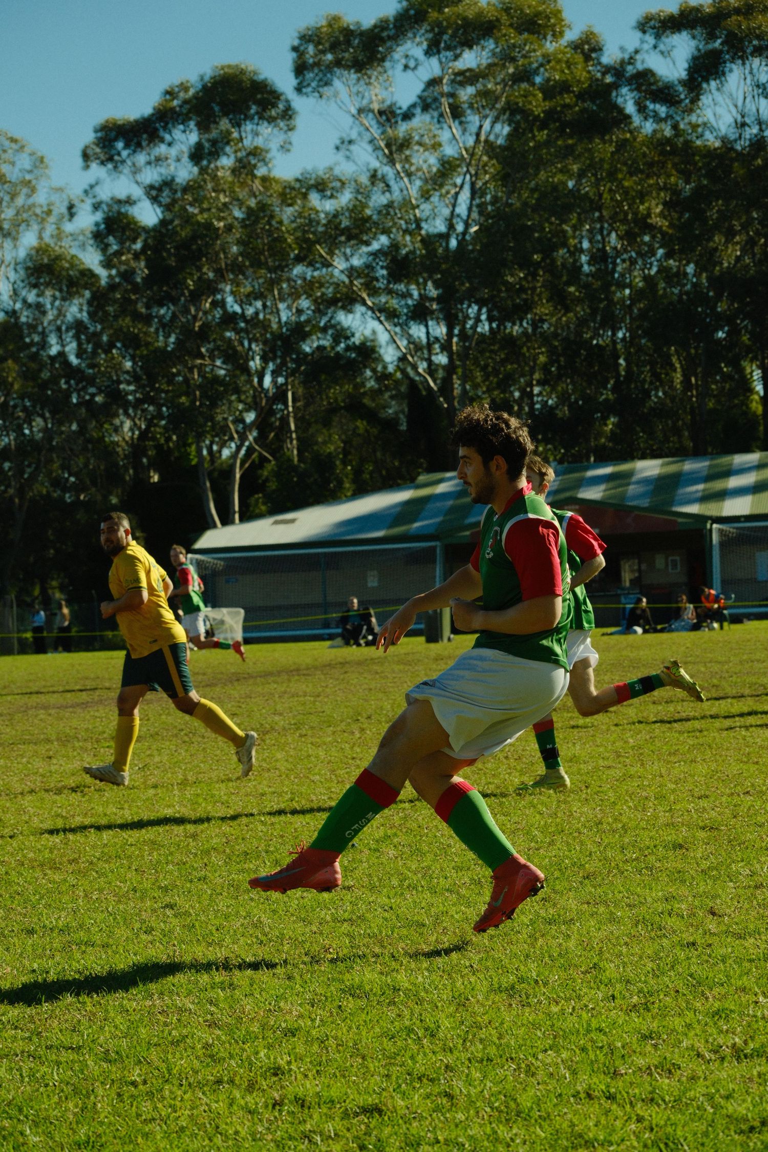 Men playing soccer on a green grass field with trees and a building in the background on a sunny day.