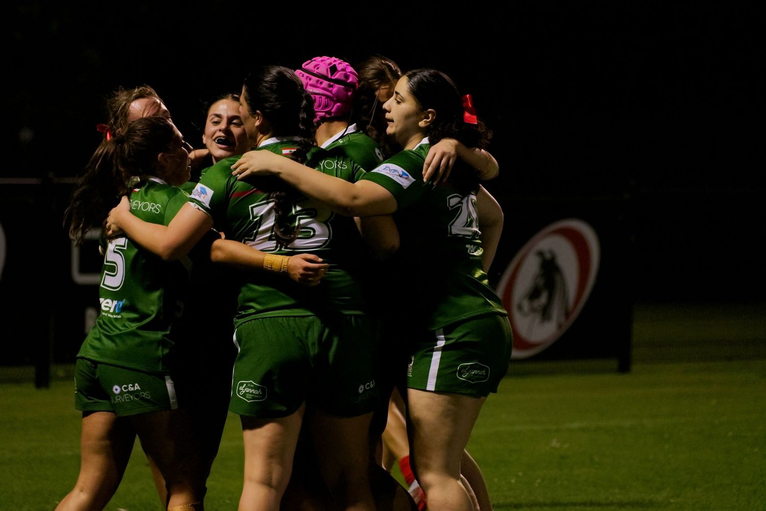 Women rugby players in green uniforms celebrating together on field at night.