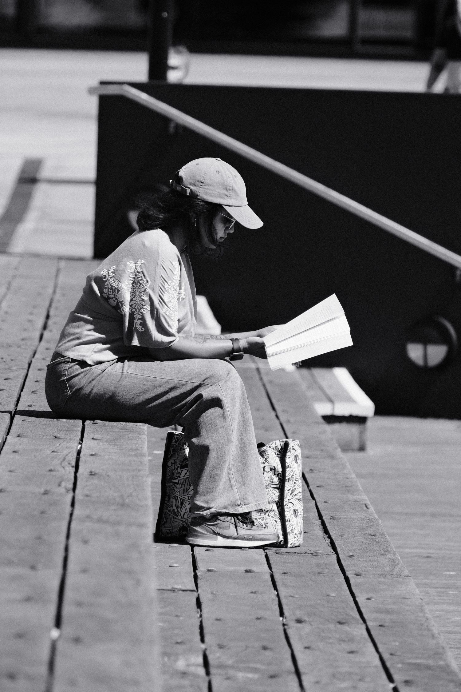 Black and white photo of a person sitting on wooden steps reading a book outdoors, wearing a cap and casual clothes.