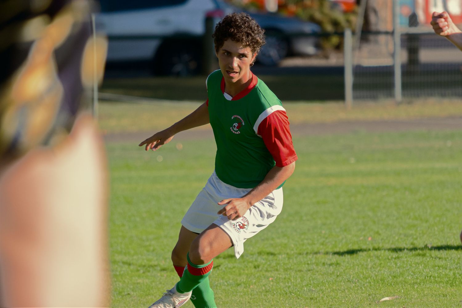 Young MSFC soccer player in green and red uniform dribbles the ball on a sunny grass field.