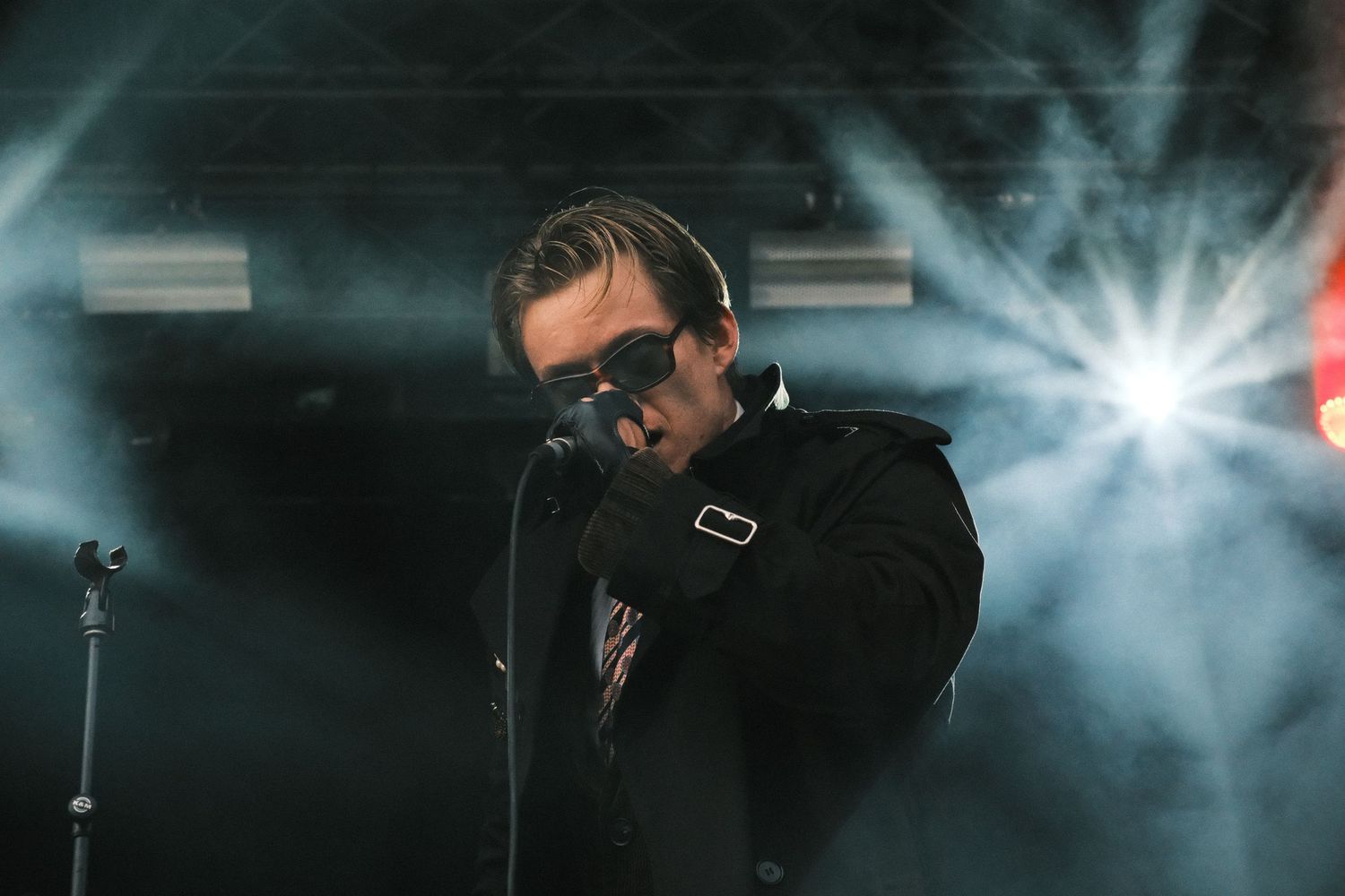 Male singer performing on stage in black suit and sunglasses, dramatic stage lighting beams in background.