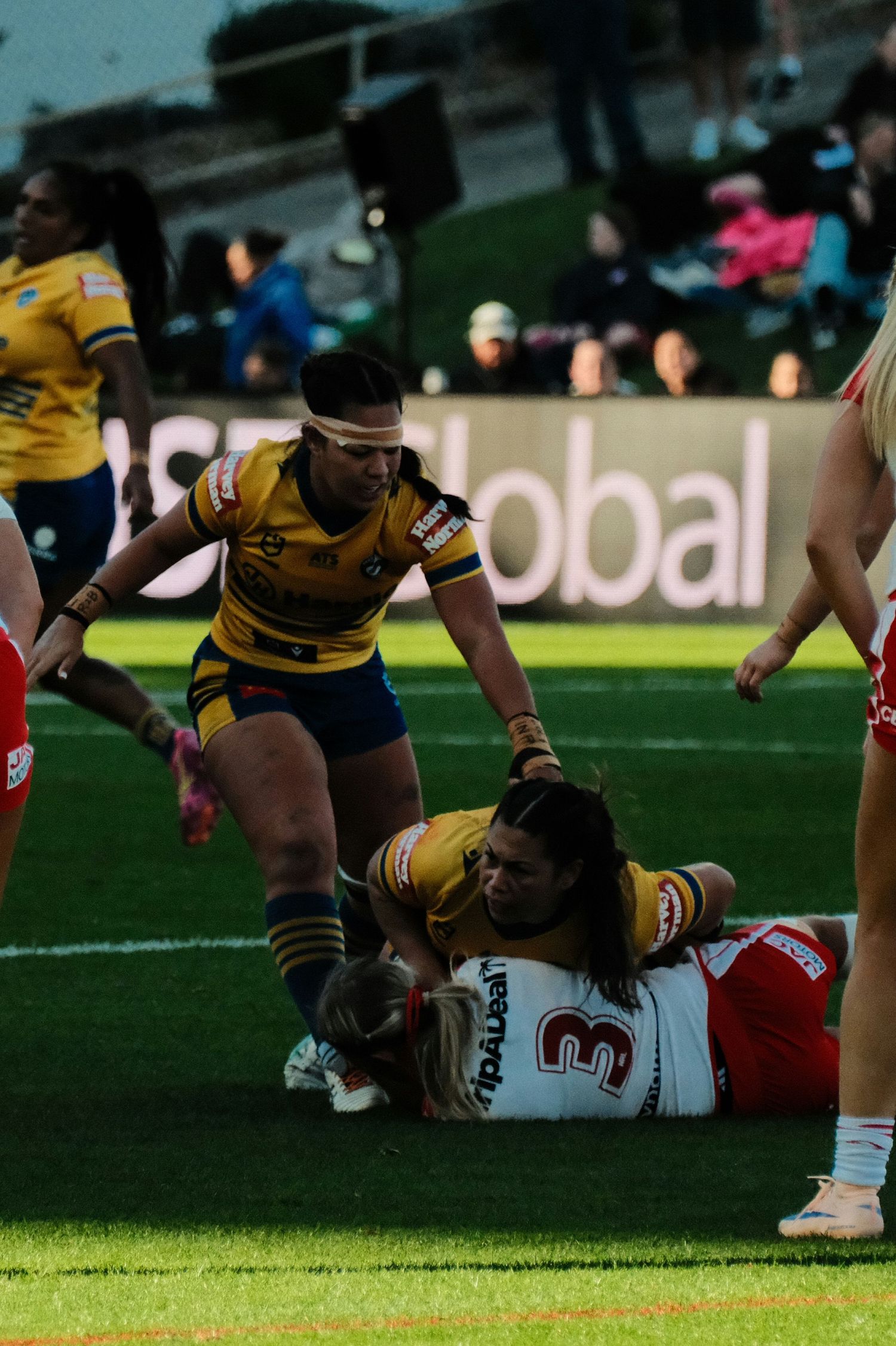 Women's rugby league players tackle during a competitive match, with players in yellow and red uniforms on a green field.