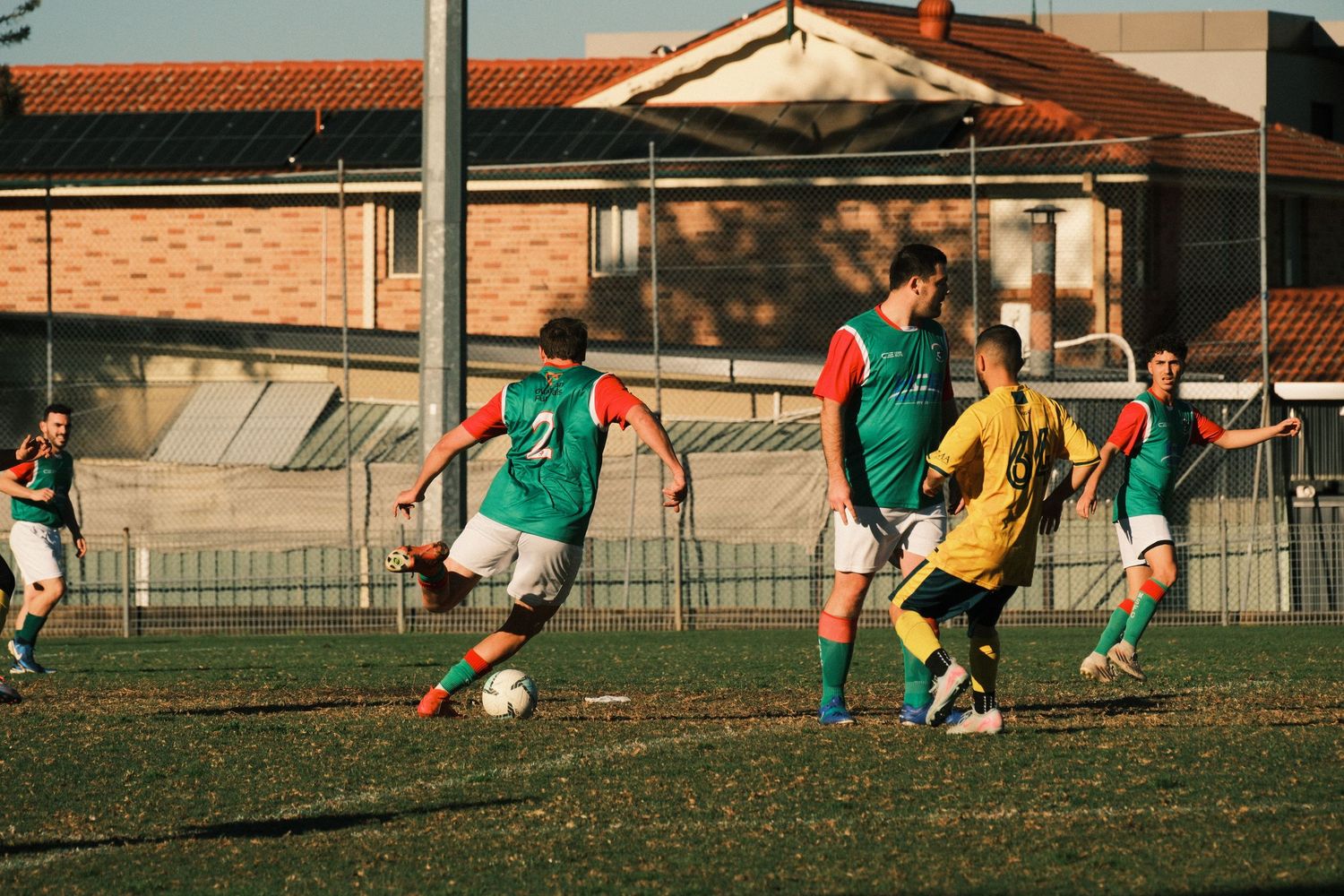Youth soccer players in green and red uniforms compete on a grass field near a brick building during a sunny match.
