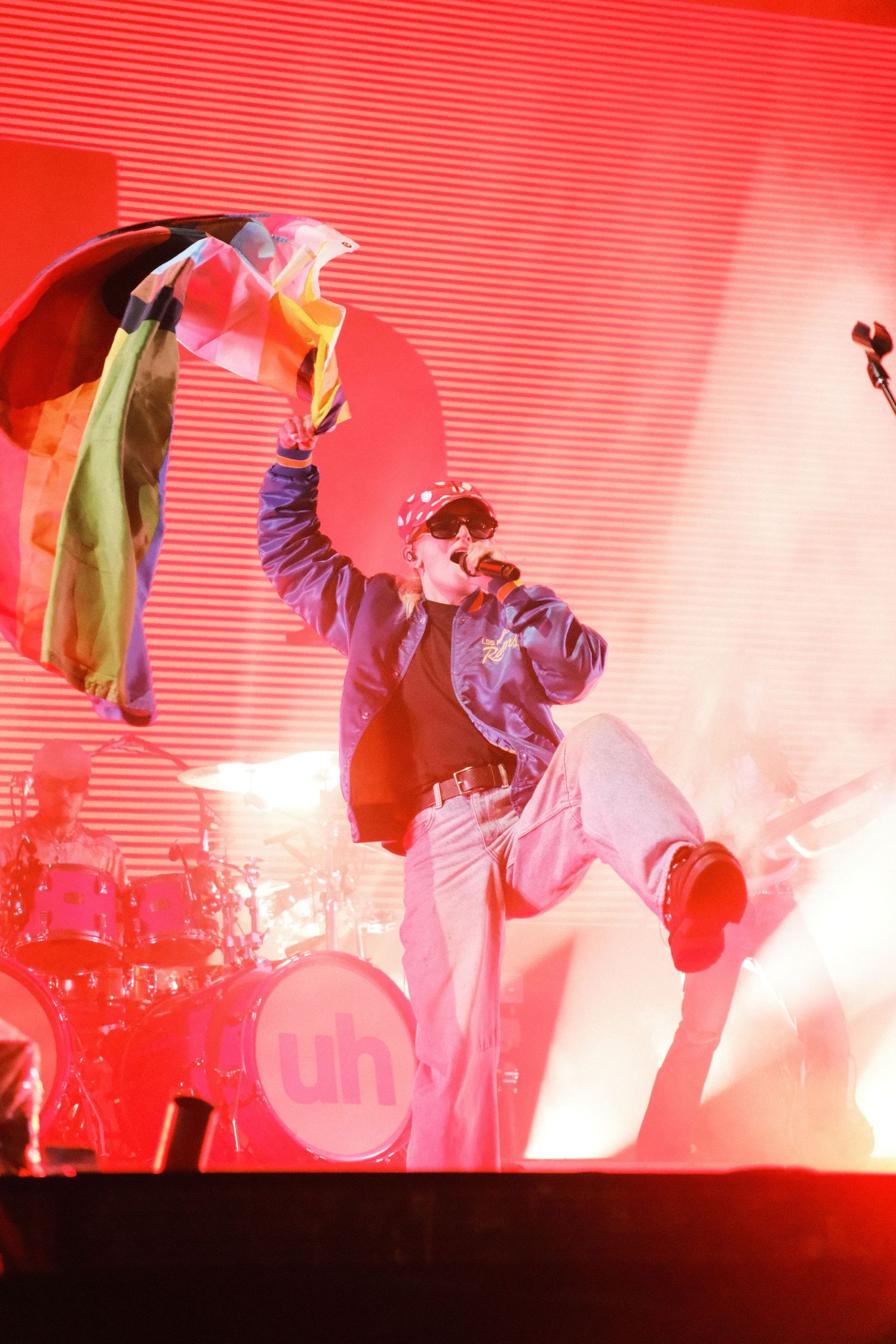 Energetic performer waves rainbow flag on red-lit stage during live concert performance.