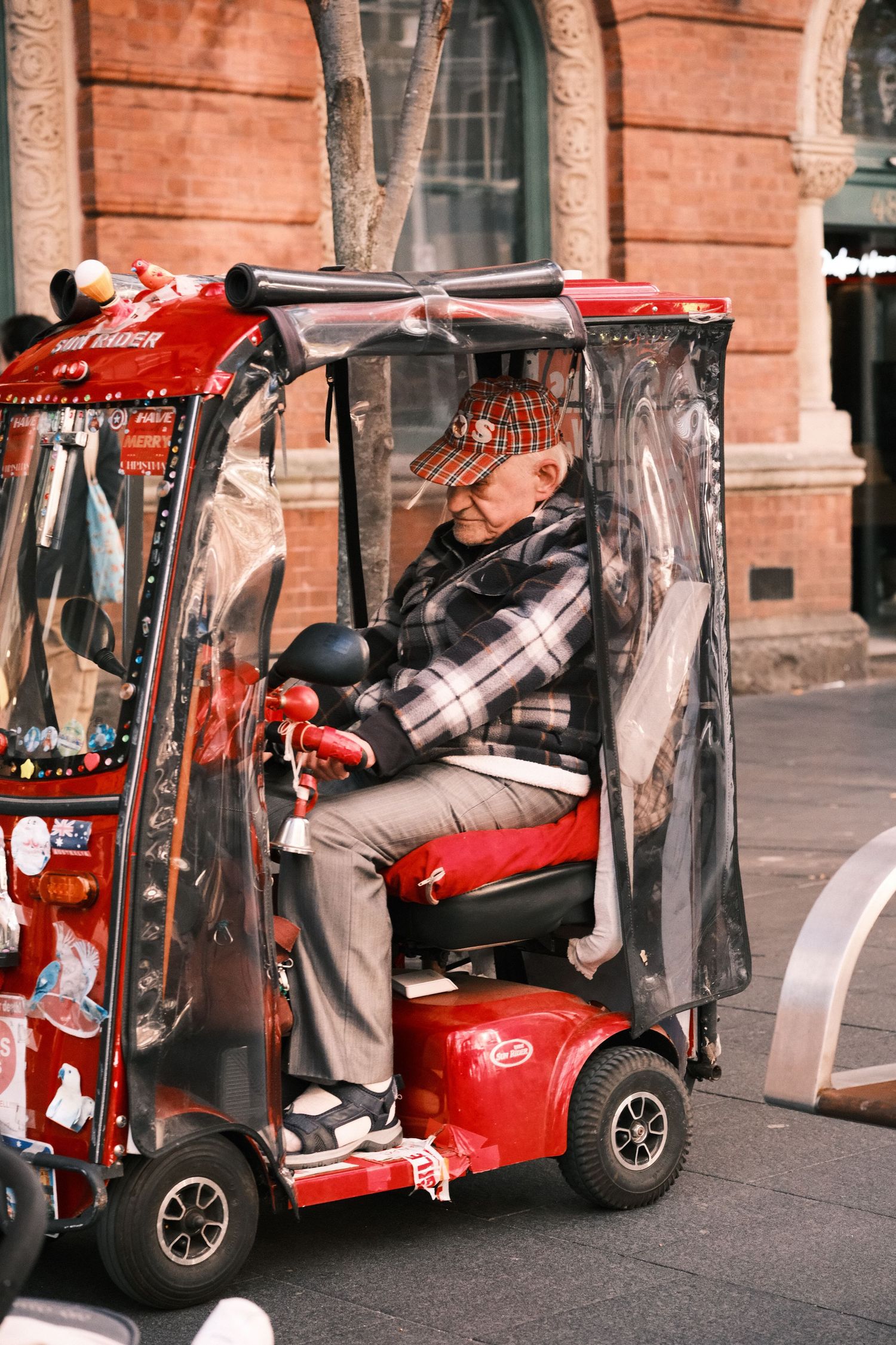 Elderly man riding a decorated red mobility scooter with a clear plastic canopy on a city street.