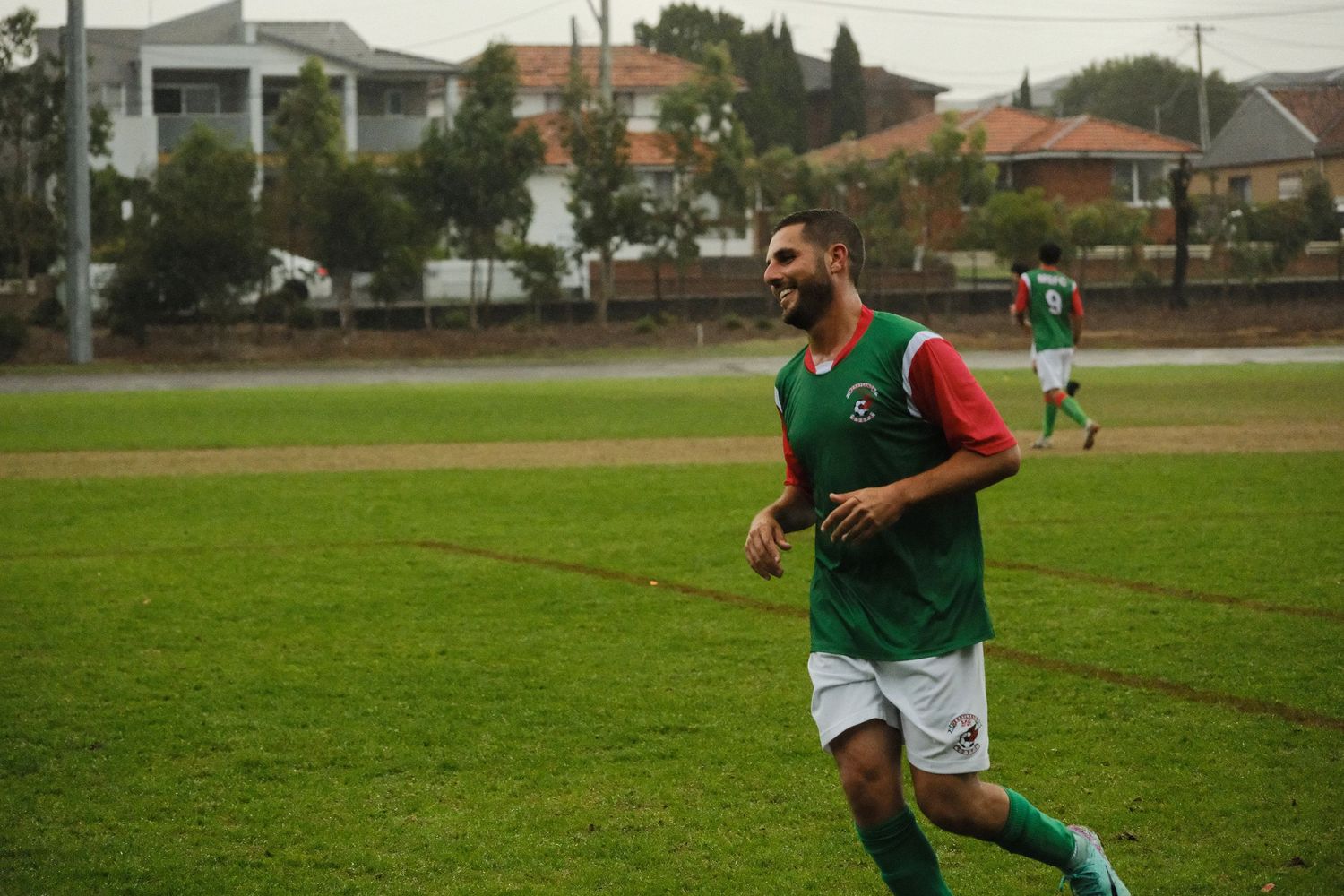 Smiling soccer player in green and red uniform jogging on a grass field outdoors.