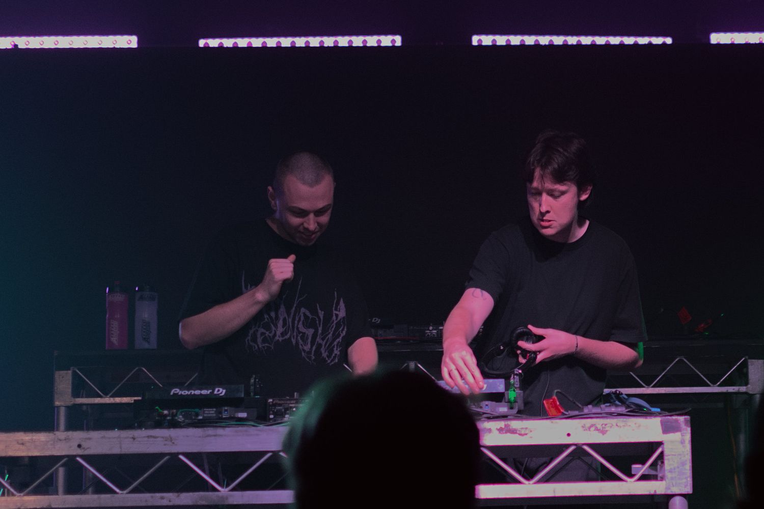 Two DJs perform behind decks on stage under purple and white lights at live music event.