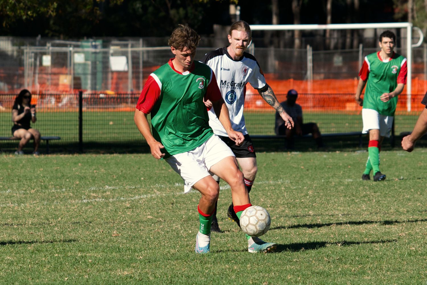 Soccer players in green and red jerseys compete for the ball during an outdoor match on a grass field.