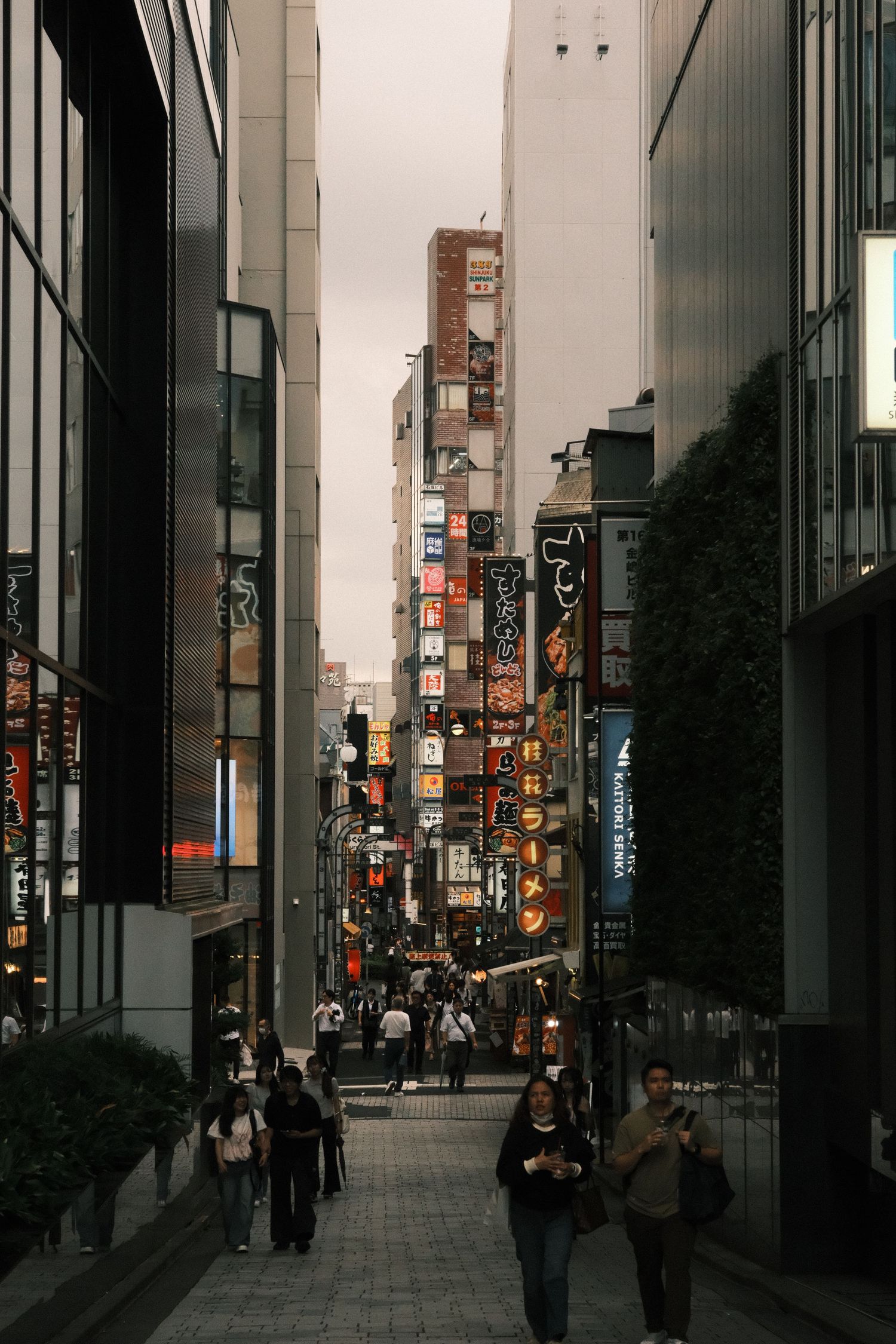 Busy urban alley in Tokyo lined with colorful neon signs and pedestrians walking between modern buildings at dusk.