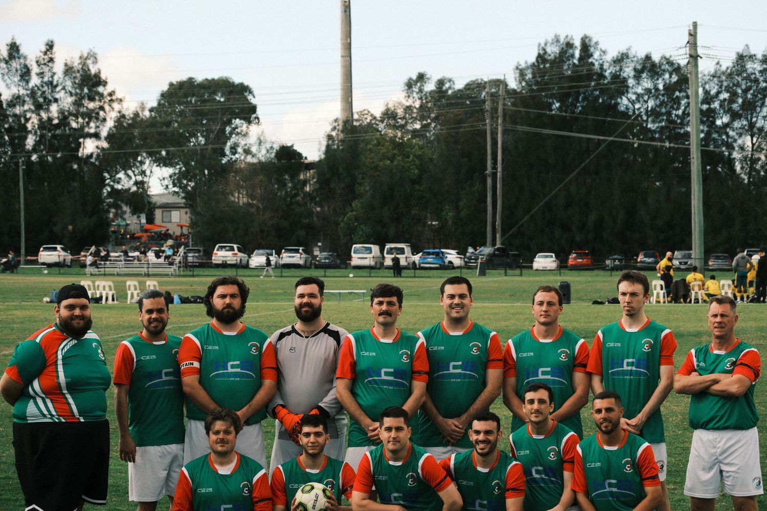 Soccer team posing in green and orange jerseys on a field, with trees and parked cars in the background.