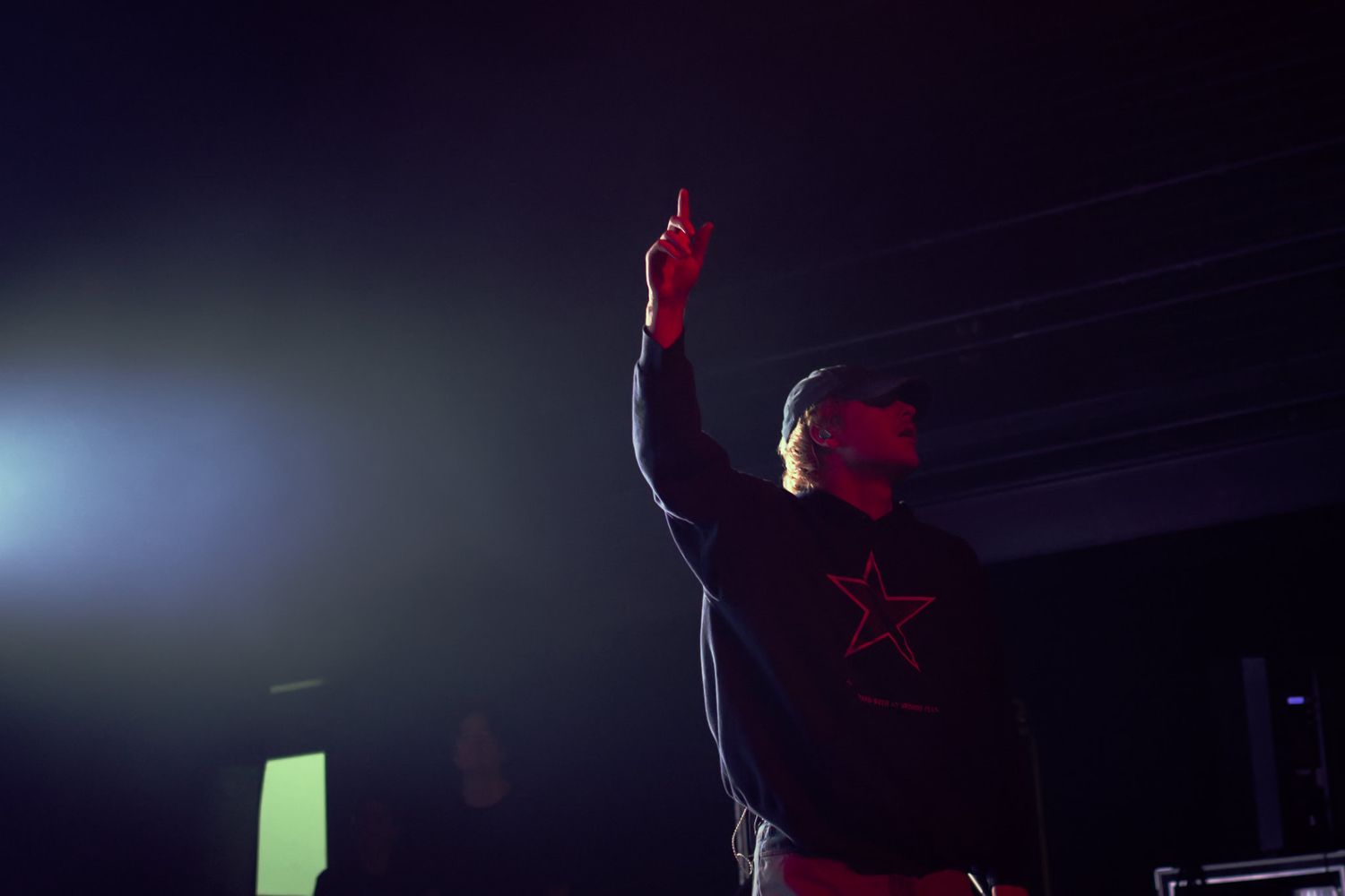 Performer raises fist on dark stage wearing black star hoodie at live hip-hop concert.