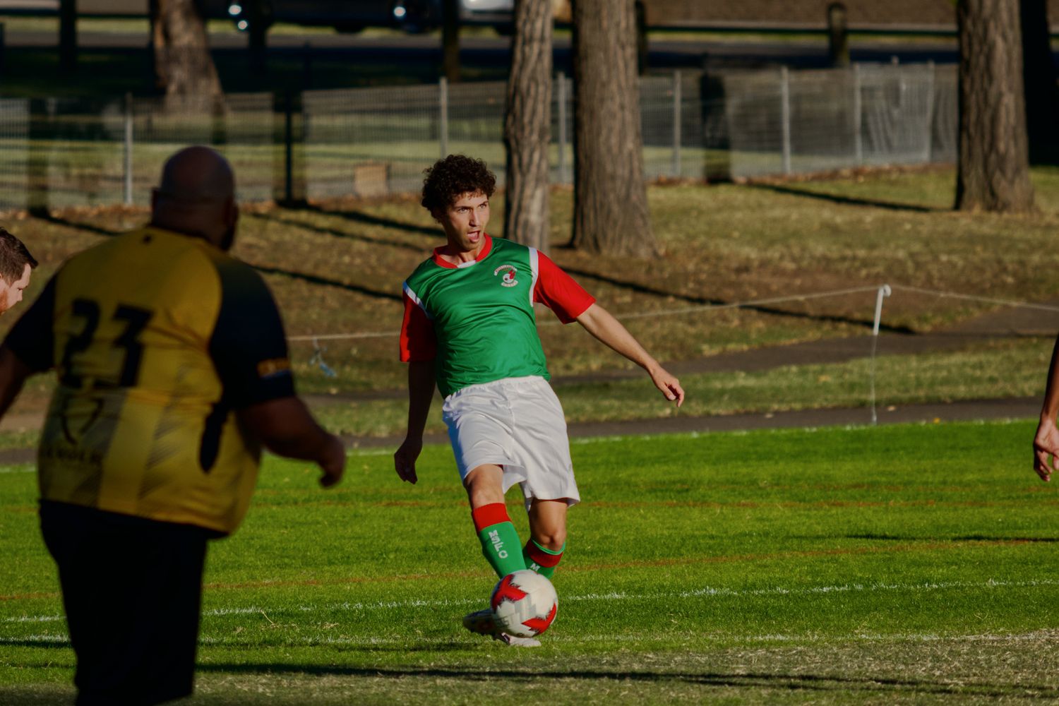 Young soccer player in red and green kit controls ball during outdoor match on grass field.
