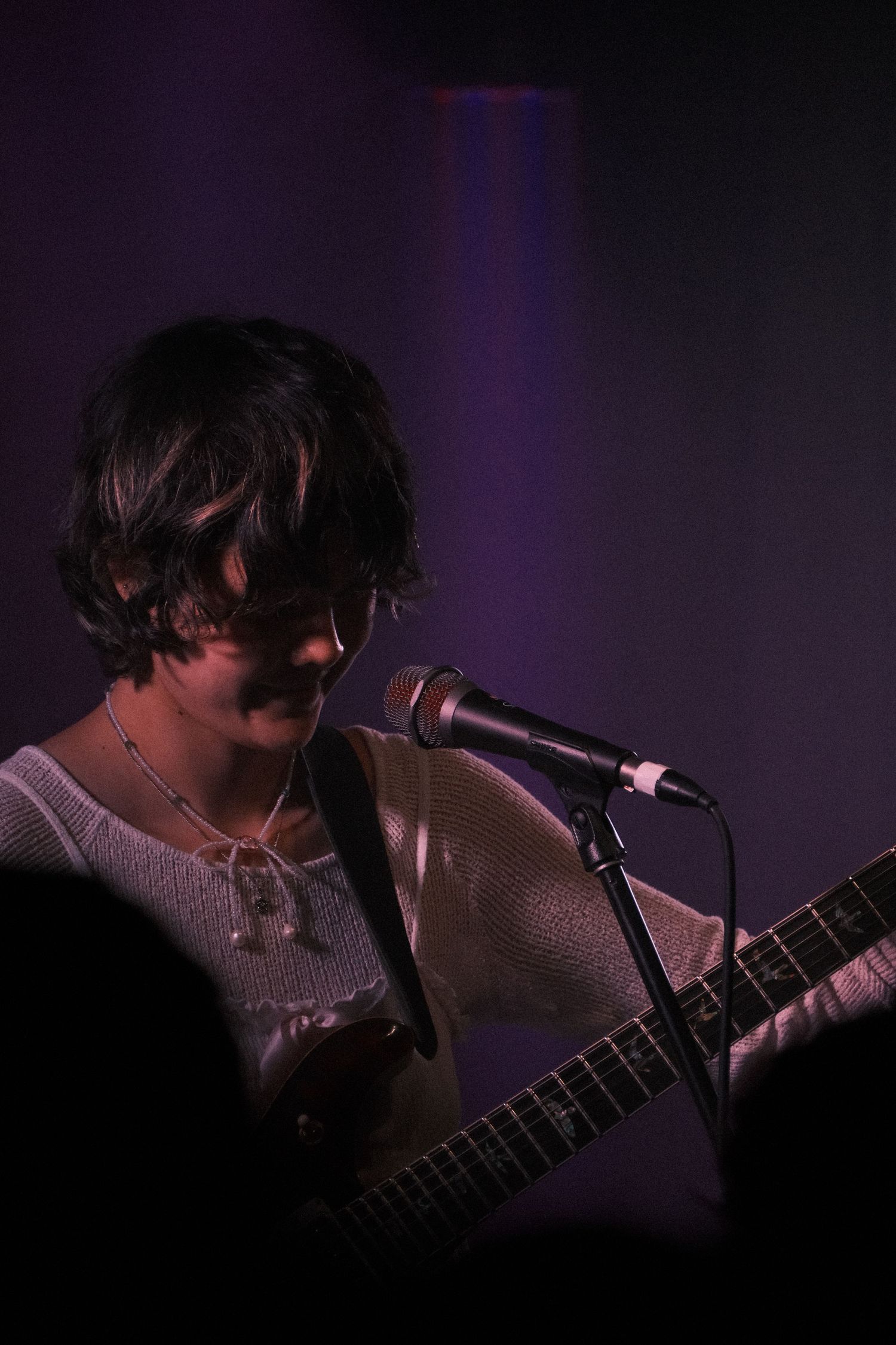 Young musician with dark hair plays electric guitar and sings into microphone on dimly lit purple-lit stage.