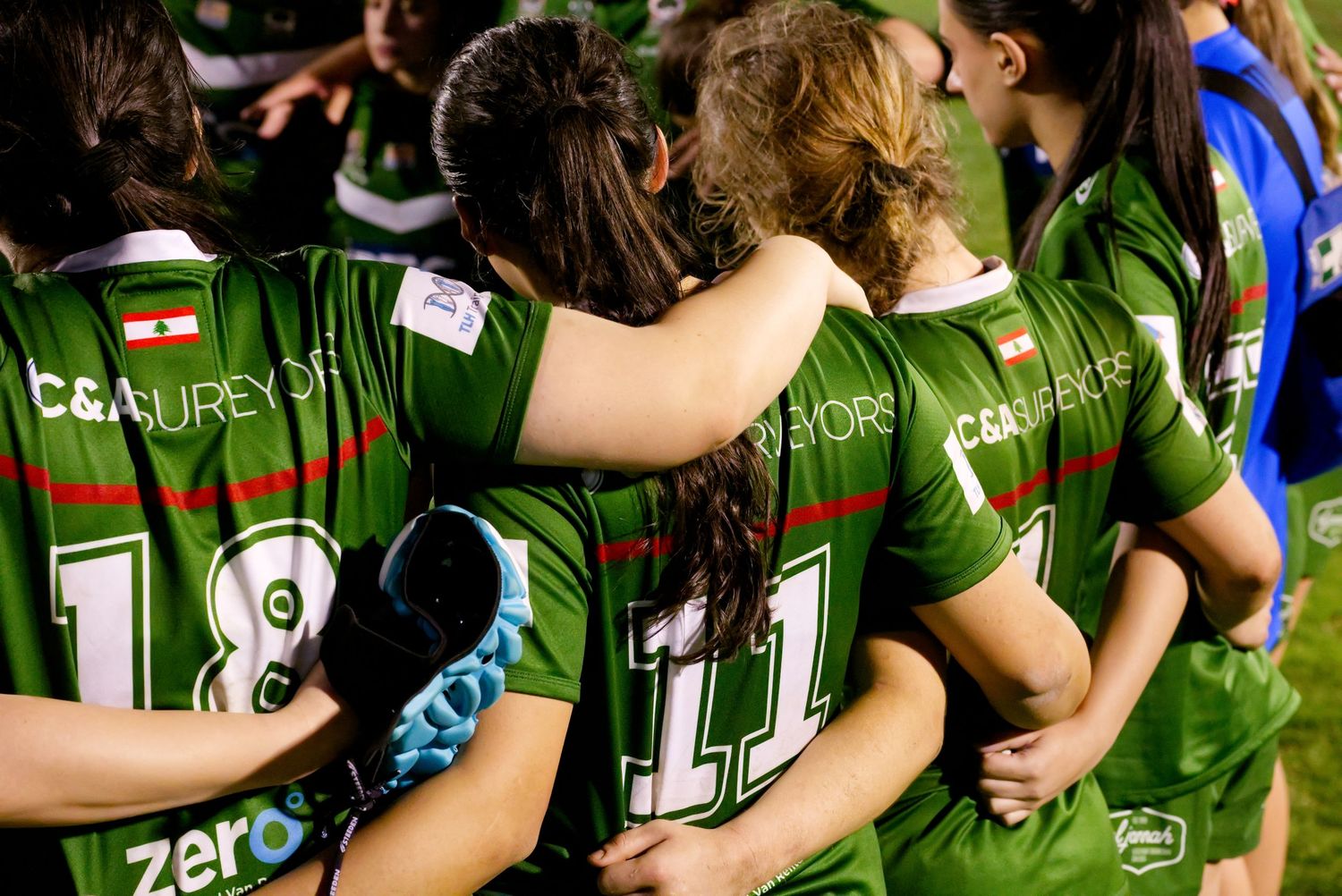 Women's rugby team in green jerseys huddle together on field during a night match.