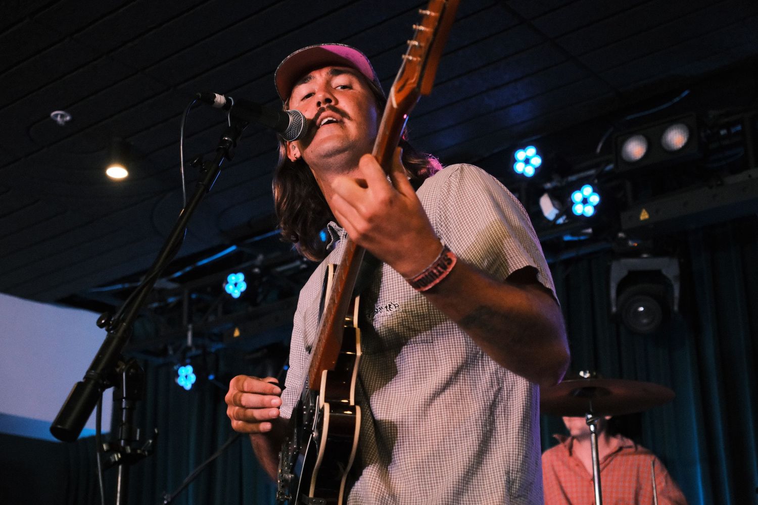 Male guitarist singing into microphone on stage at live music venue, wearing cap and striped shirt.