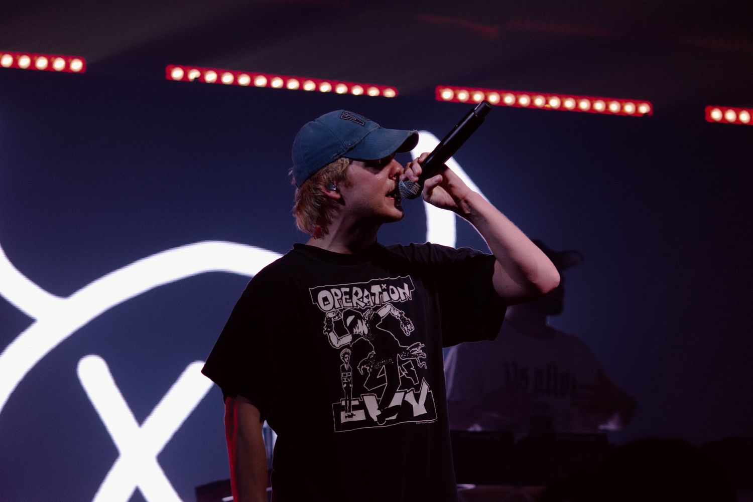Performer in blue cap and black graphic tee sings into microphone on stage with LED light display.