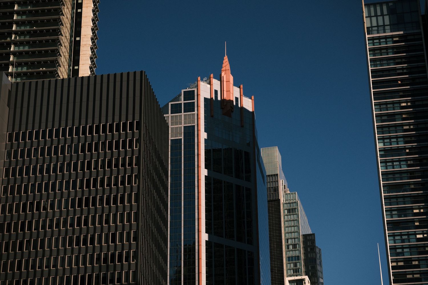 Sunlit Art Deco skyscraper crown rises above modern glass towers against a clear blue sky in a dense urban cityscape.