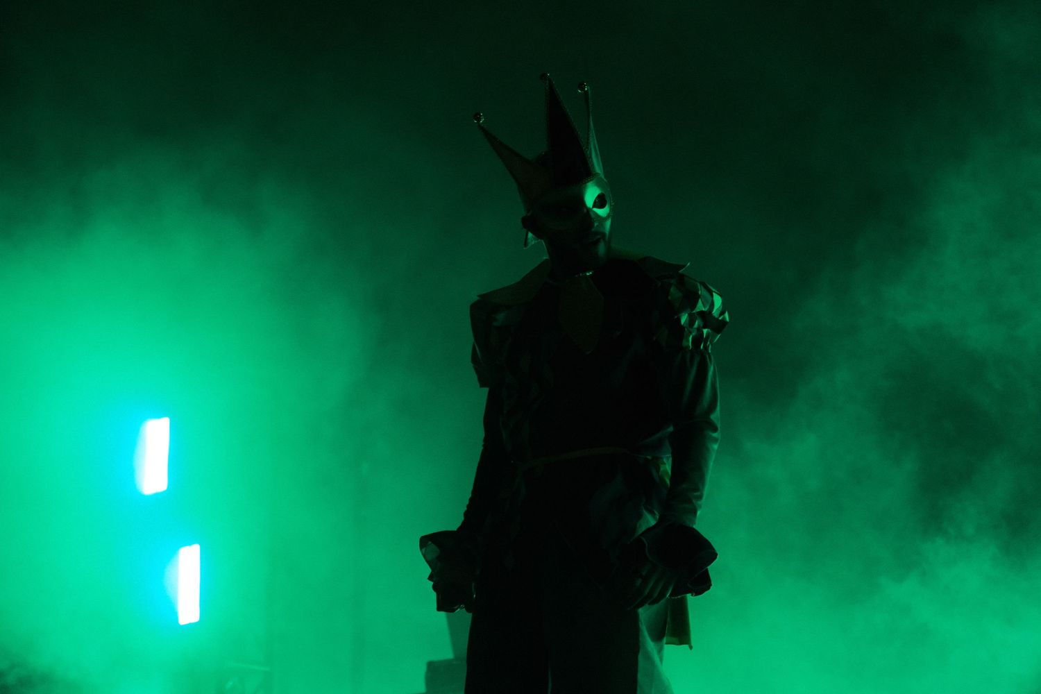 Mysterious figure in horned costume stands dramatically in green smoky stage lighting.