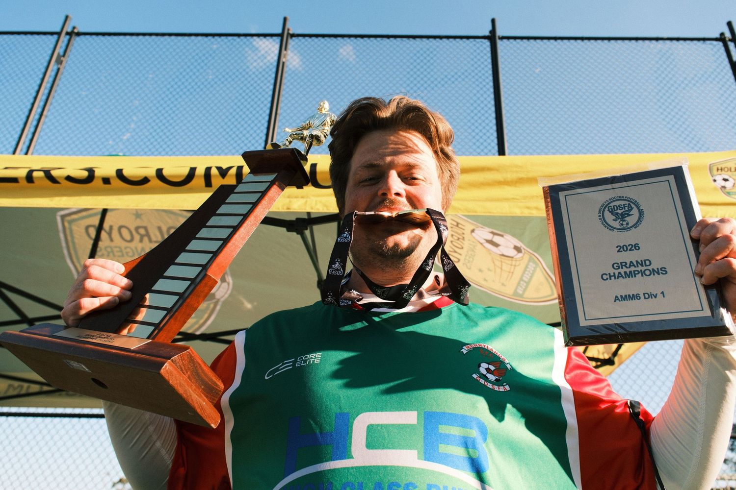 Man proudly holding Grand Champions trophy plaque and award outdoors at a sports tournament.