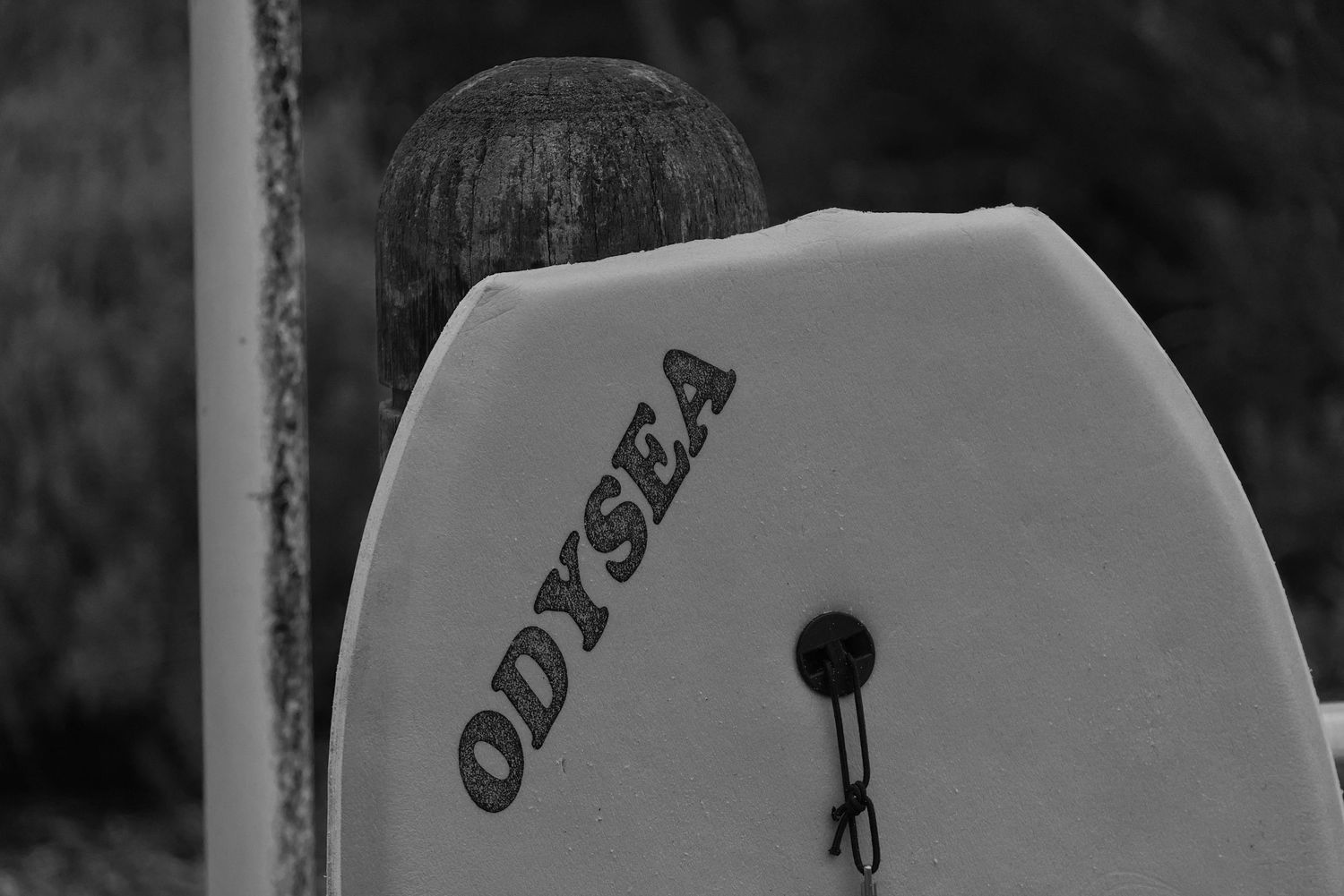 Black and white photo of an Odysea branded surfboard leaning against a post.