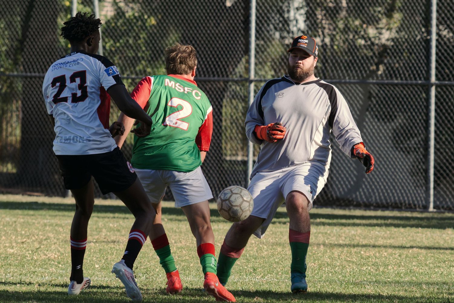 Three soccer players compete for the ball during an outdoor match, including a goalkeeper in gray.