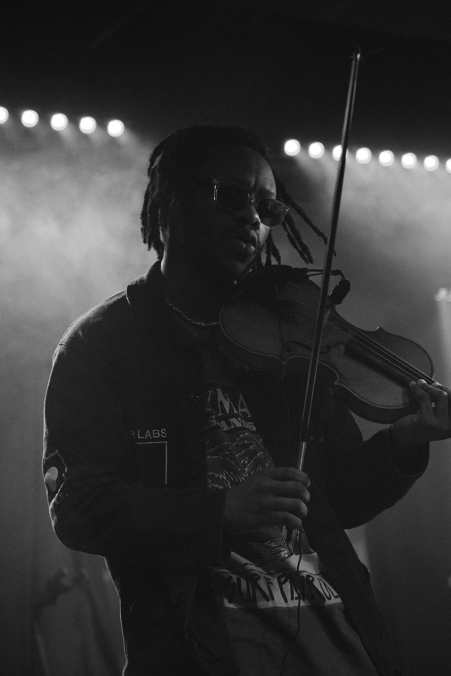 Black and white photo of a musician with dreadlocks playing violin on a smoky stage under bright lights.