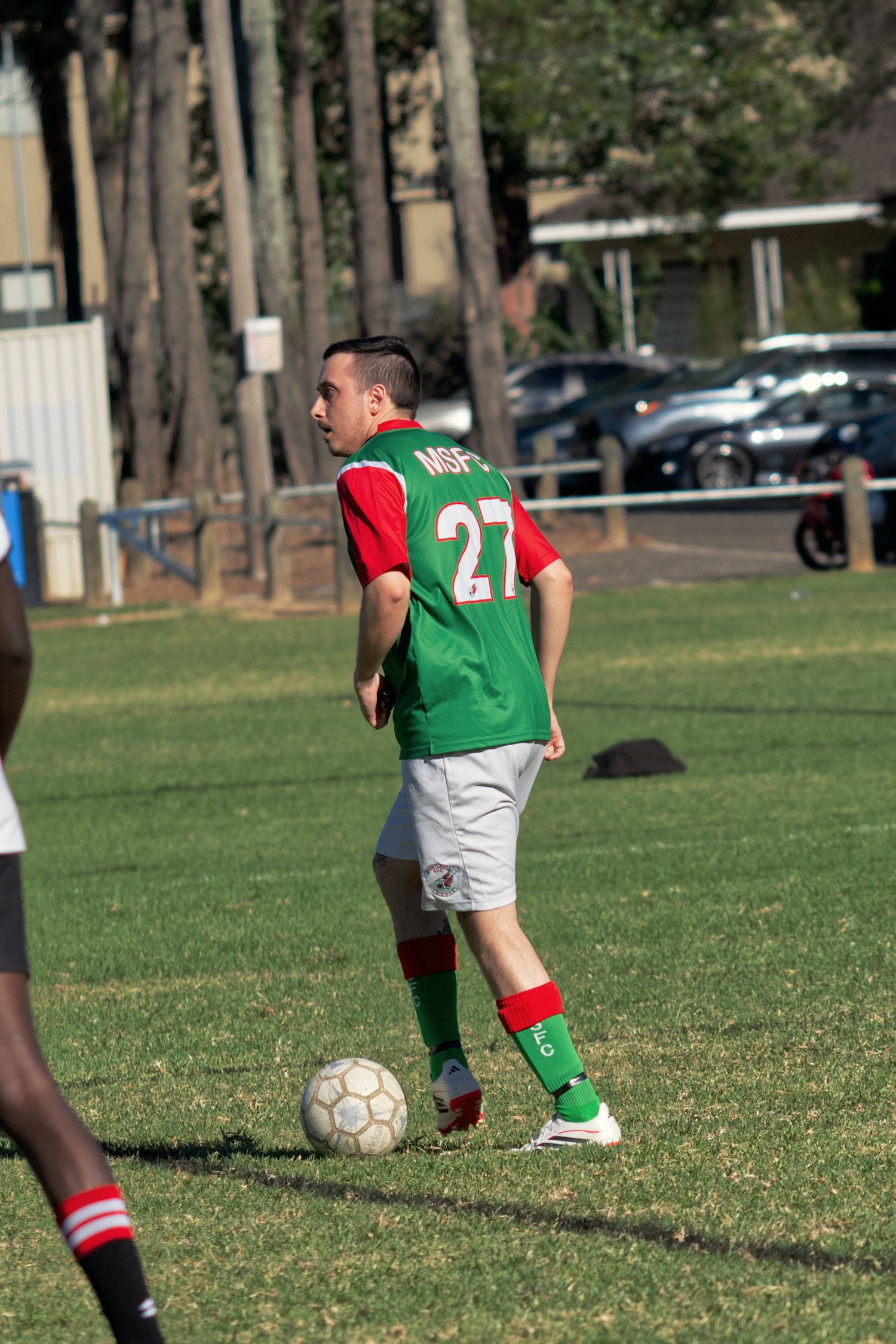 Soccer player wearing number 27 green MSFC jersey dribbles a ball on a sunny grass field.