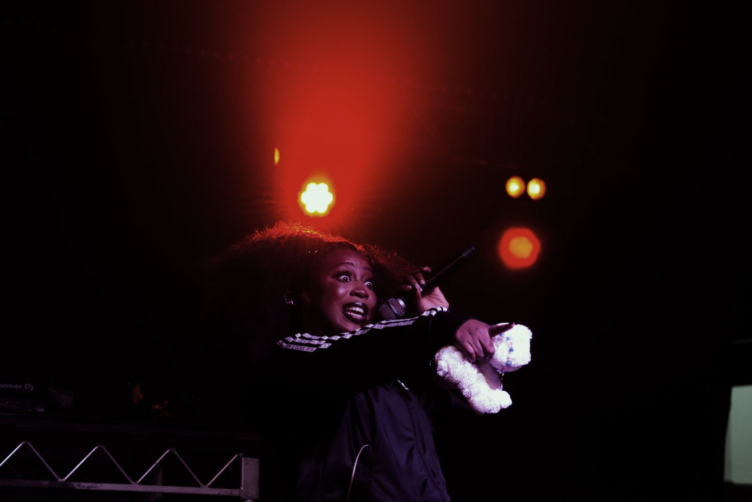 Performer in checkered outfit holds stuffed animal on stage under dramatic red and purple lighting.