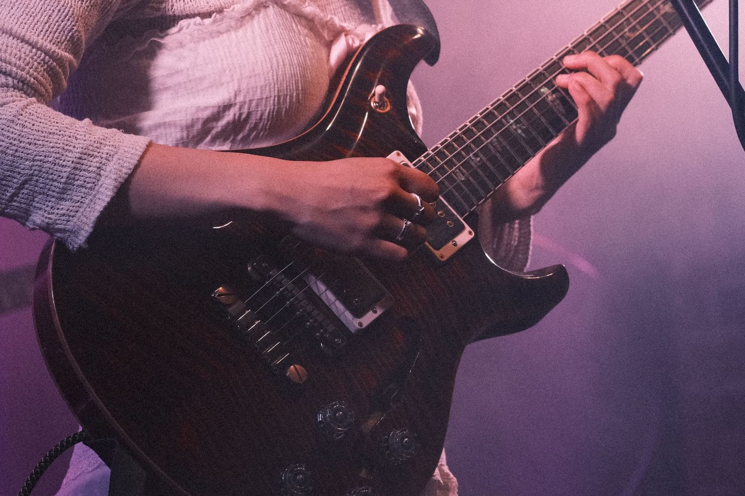 Close-up of a guitarist playing a dark electric guitar on stage under purple stage lighting.